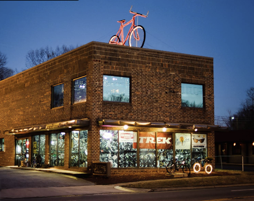 Brick bicycle shop with large windows displaying bikes inside, illuminated signs outside, and a large pink bicycle sculpture on the roof at dusk.