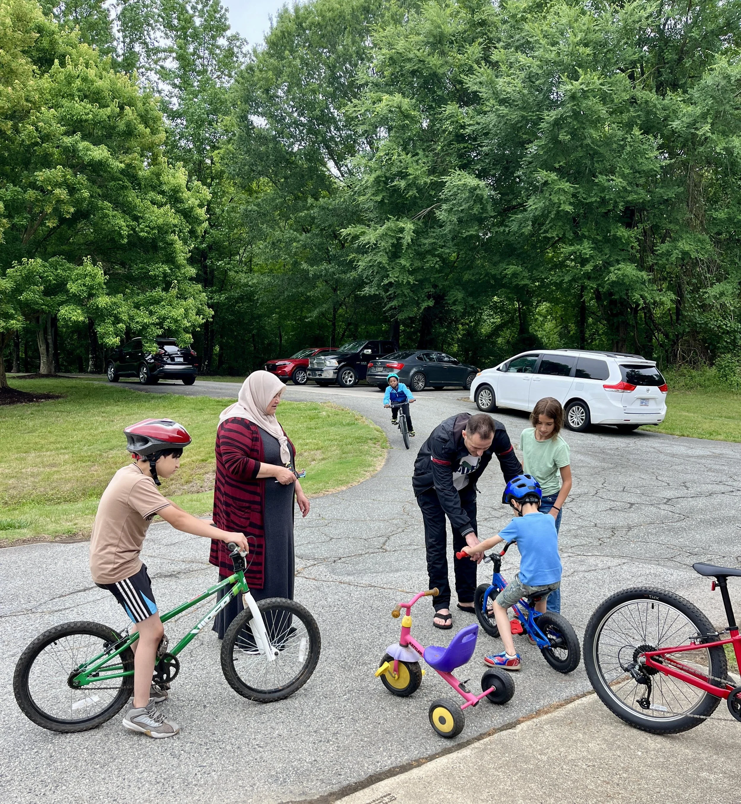 Group of children and adults with bikes at a park near a parking lot and green trees, with some children wearing helmets.