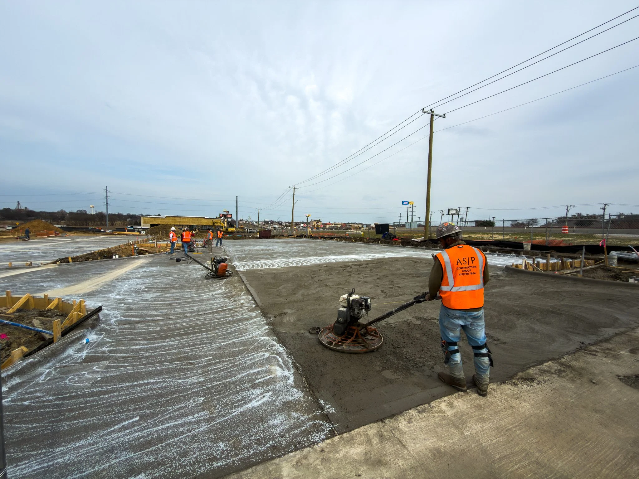 Construction workers working on a large concrete slab at a construction site with power lines and distant commercial signs visible.