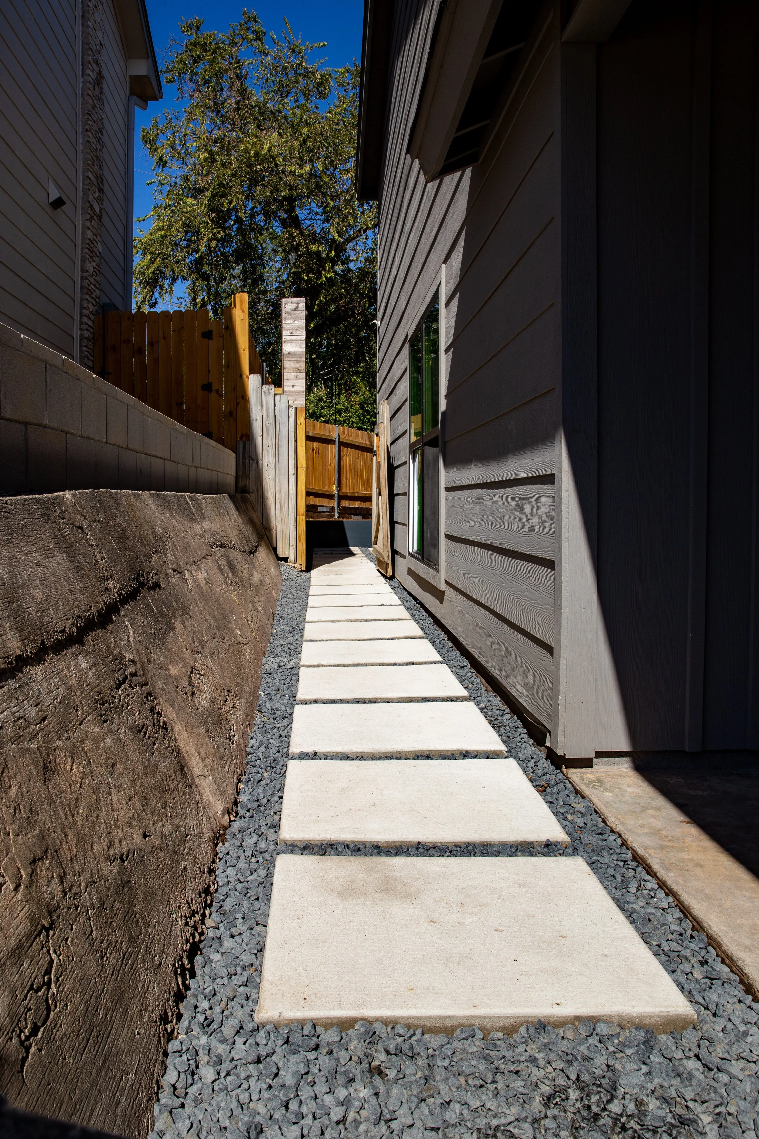 A narrow outdoor pathway with rectangular concrete stepping stones surrounded by dark gray gravel, running alongside a gray house with wooden siding on the right, and a wooden fence on the left, with green trees and a clear blue sky in the background