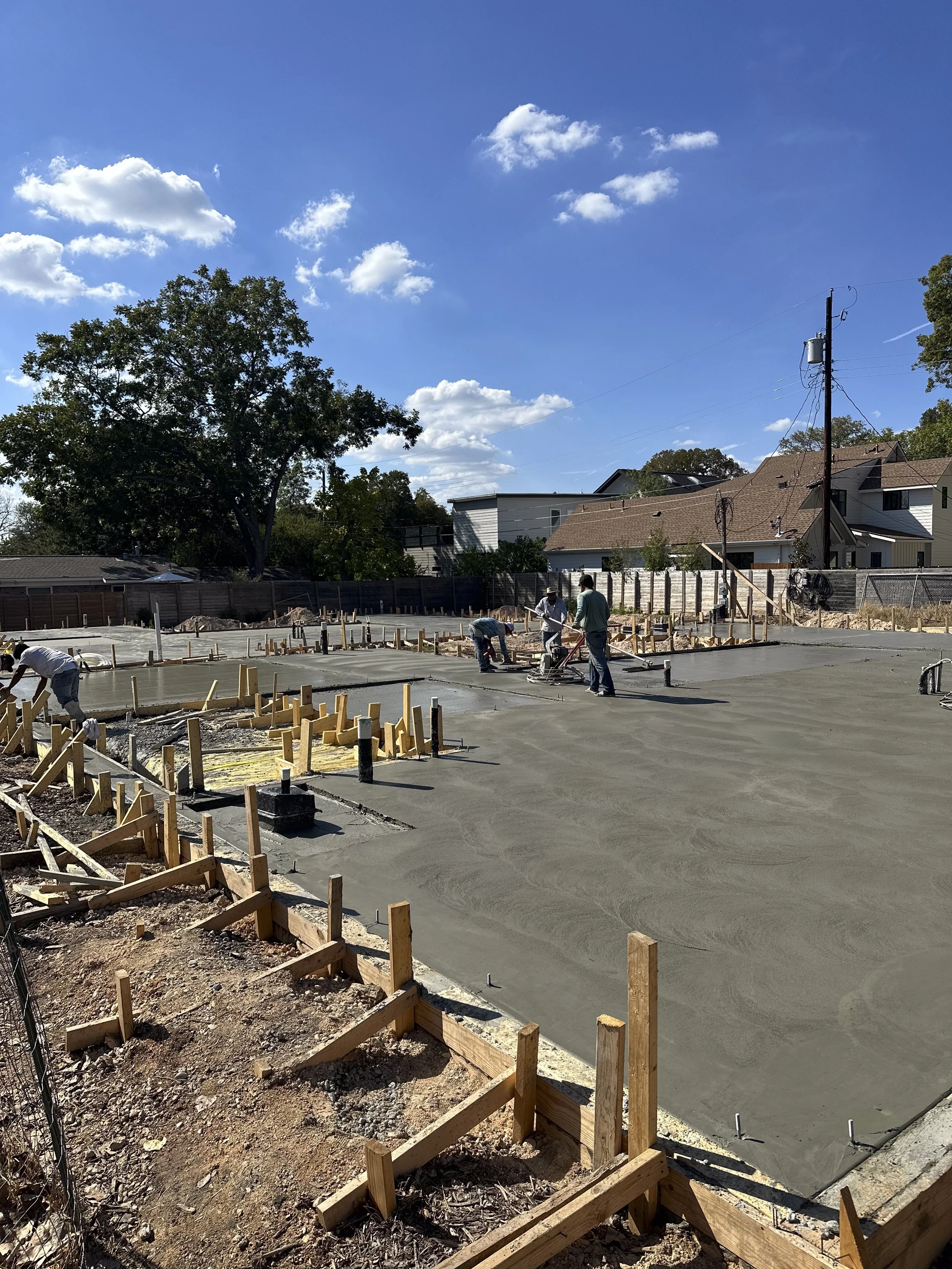 Construction site with workers pouring and finishing a concrete foundation, wooden forms in place, residential houses and trees under a blue sky with scattered clouds.