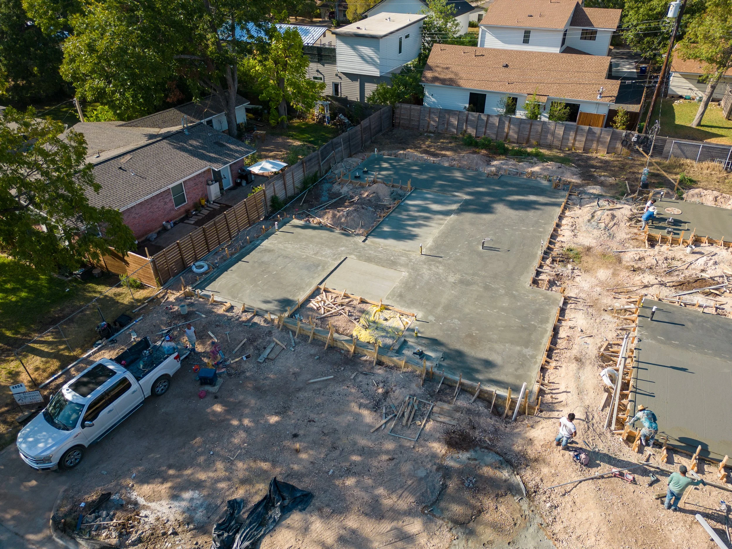 Aerial view of a construction site with a concrete foundation and wooden framework, surrounded by residential houses and trees.