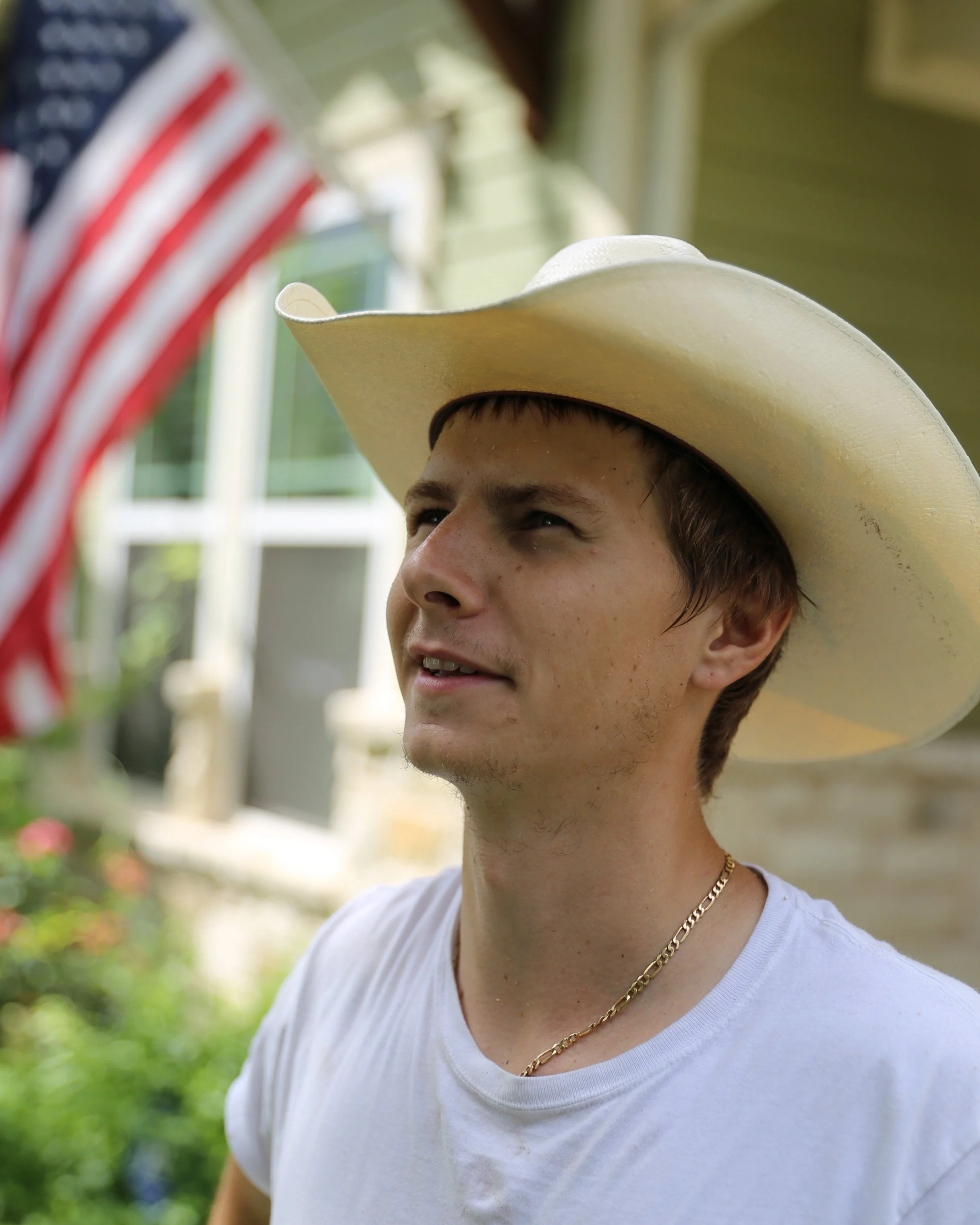 Close-up of a young man wearing a white cowboy hat, a white T-shirt, and a gold chain, standing outdoors with greenery and a house in the background.
