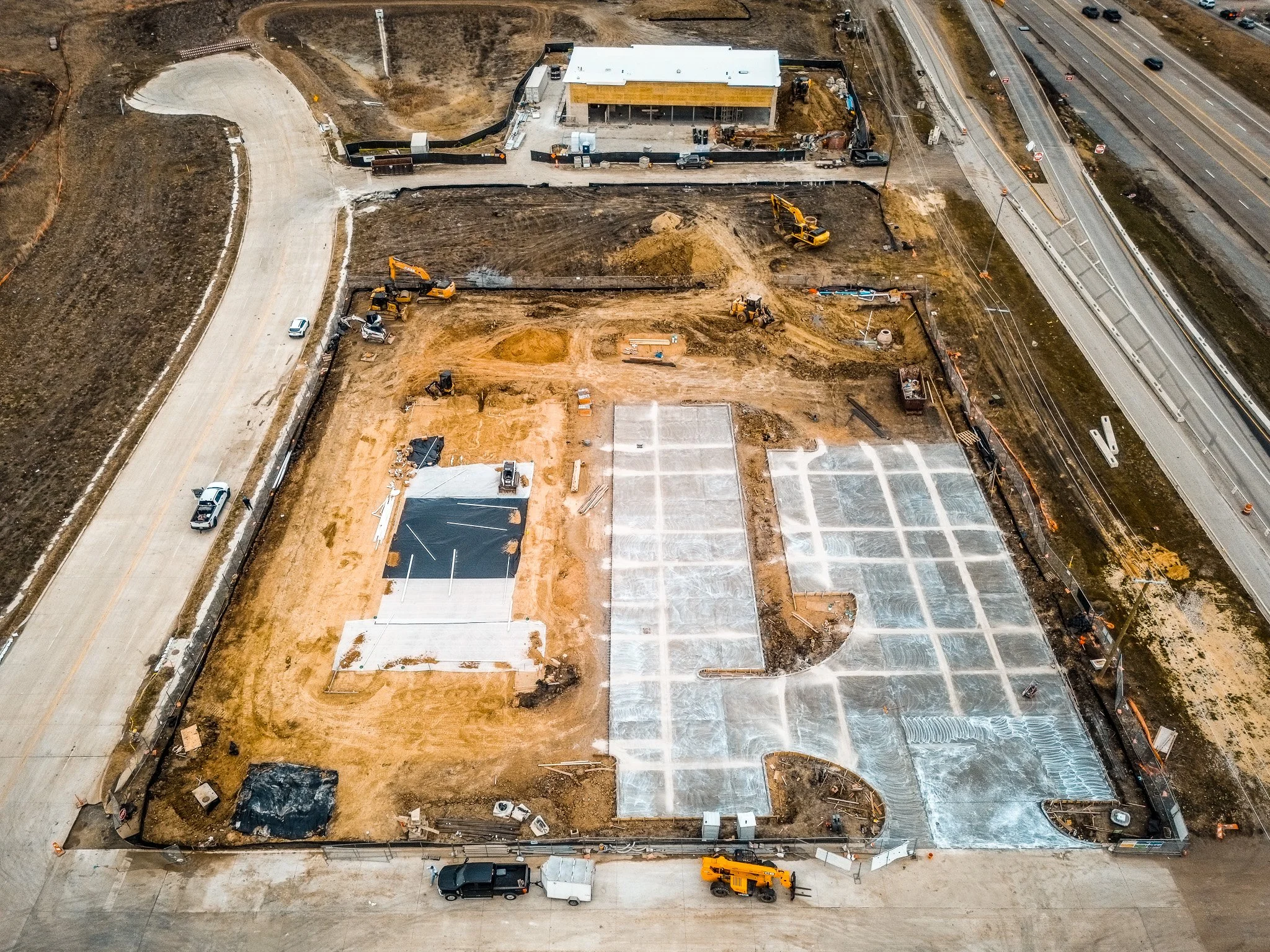 Aerial view of a construction site with machinery, partially completed concrete areas, and a nearby highway with traffic.