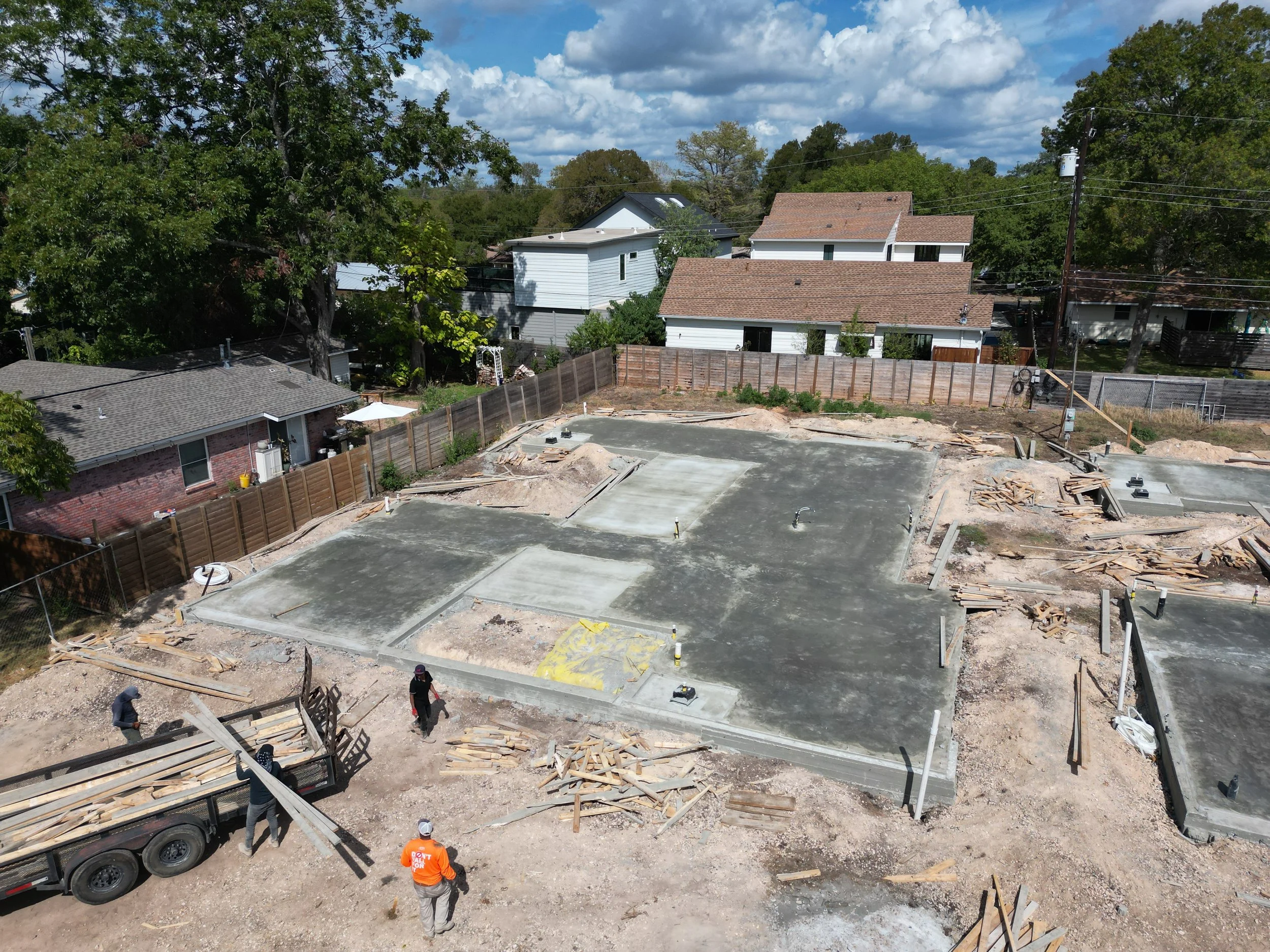 Construction site with foundations poured for new buildings, workers moving wood and construction materials, and residential houses in background with trees and a partly cloudy sky.