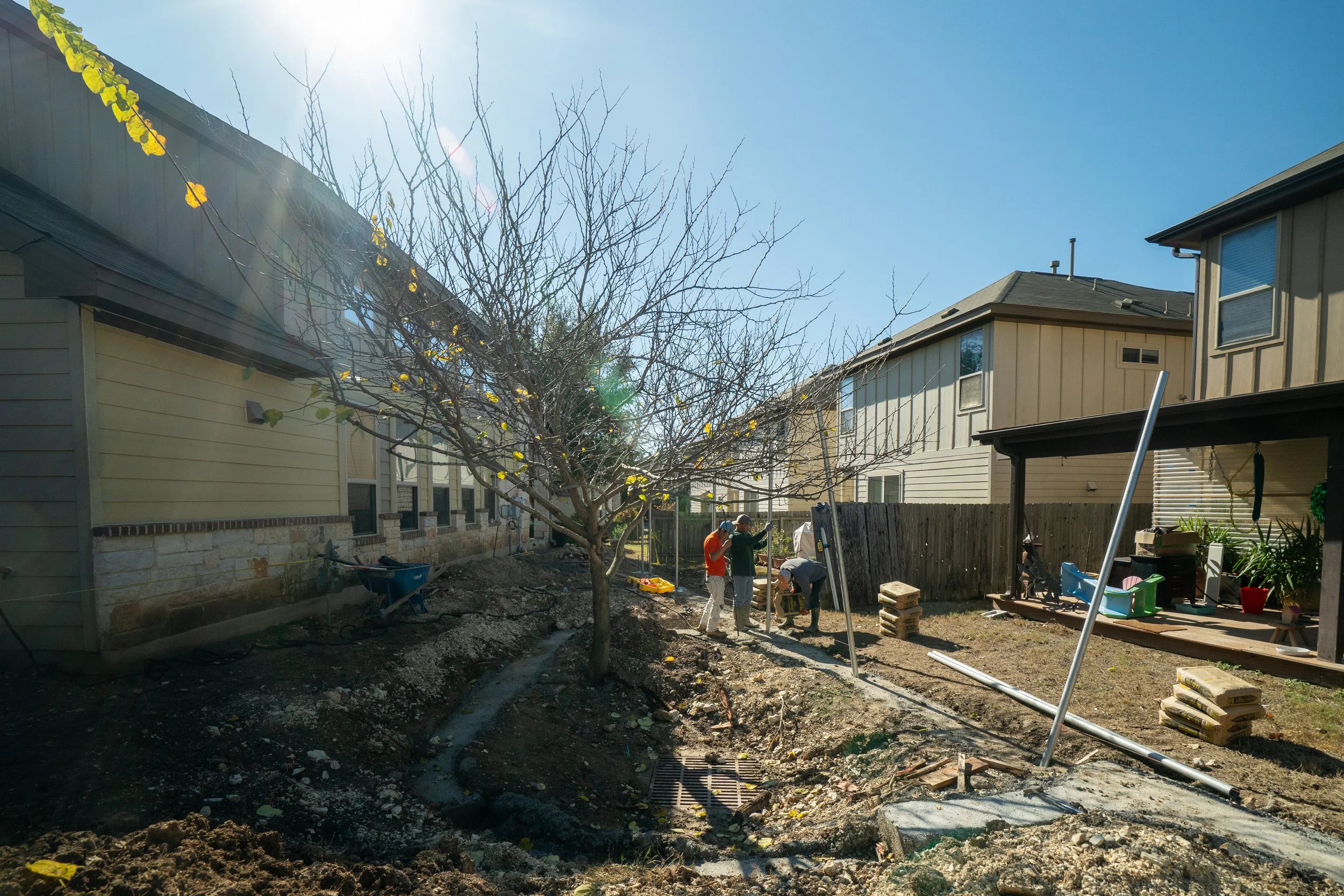 Backyard renovation with workers among gardening tools, construction materials, and a leafless tree under a bright blue sky.
