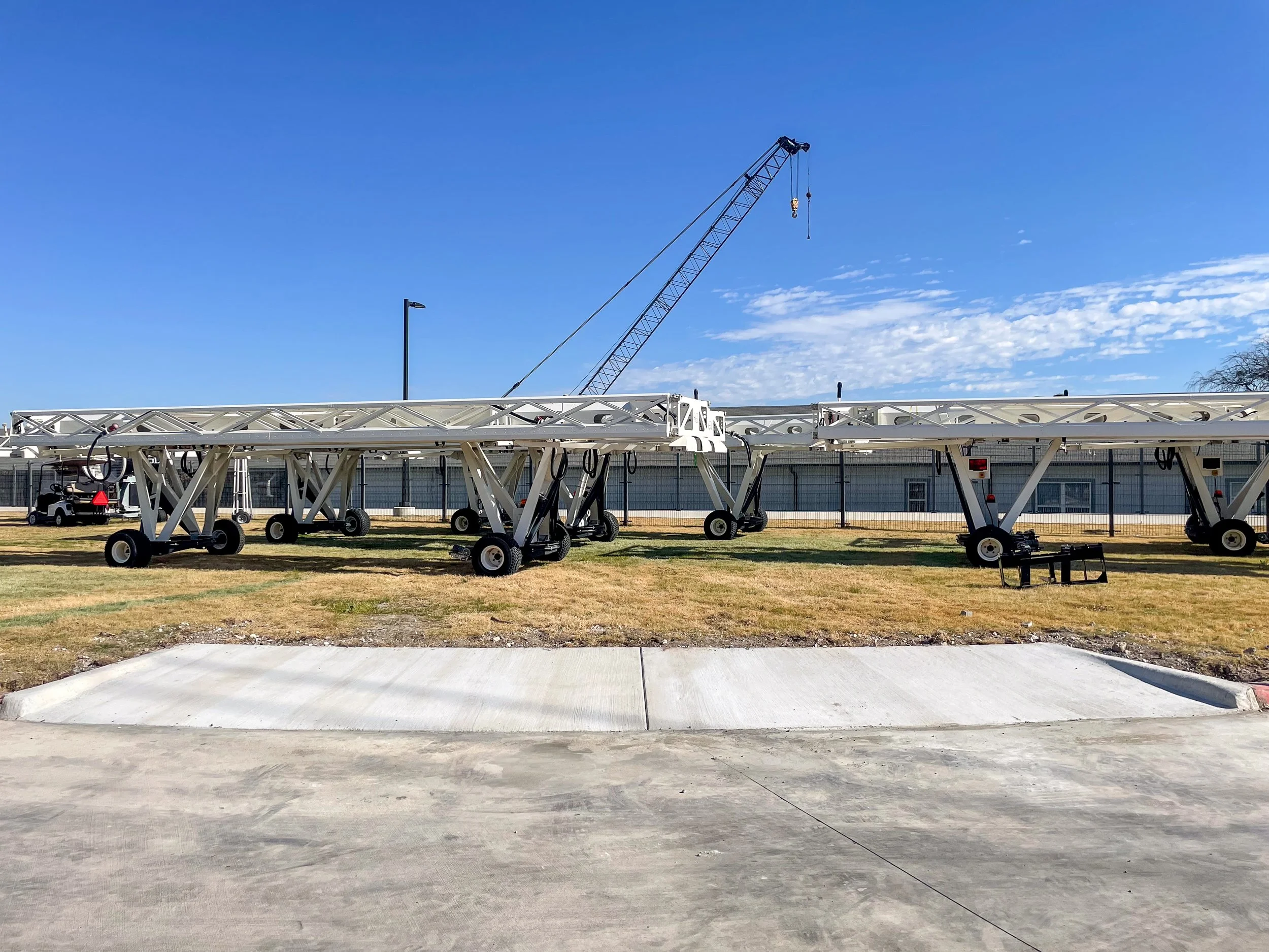 A construction crane in a clear blue sky, with a white, heavy-duty base structure on a patch of grass and concrete surface.