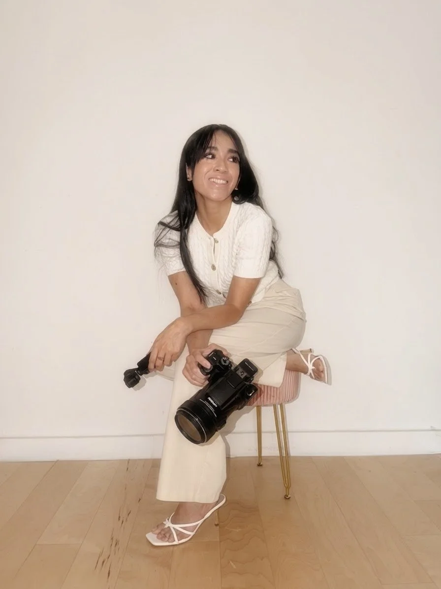 A woman with long black hair sitting on a pink and gold stool against a plain white wall, smiling while holding a camera in her right hand.