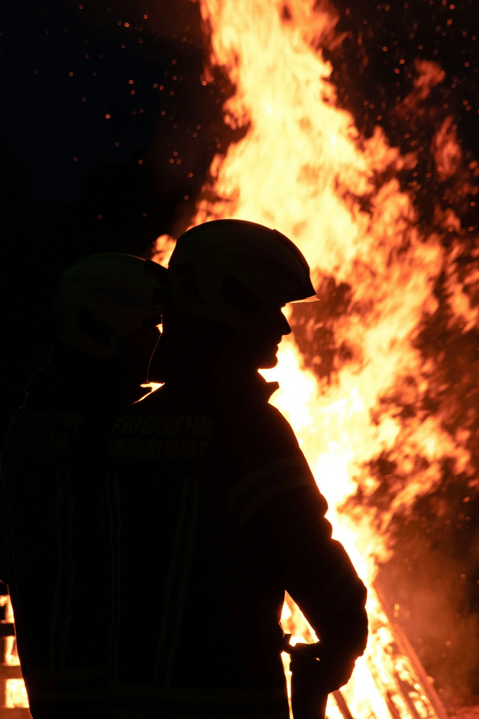 Silhouette of a firefighter wearing a helmet during a large fire with flames and smoke in the background.