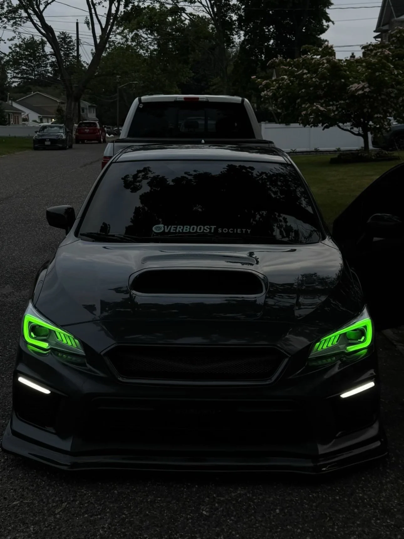 Black sports car with green LED headlights parked outdoors, with custom hood scoop and window decal reading "VERBOOST SOCIETY".
