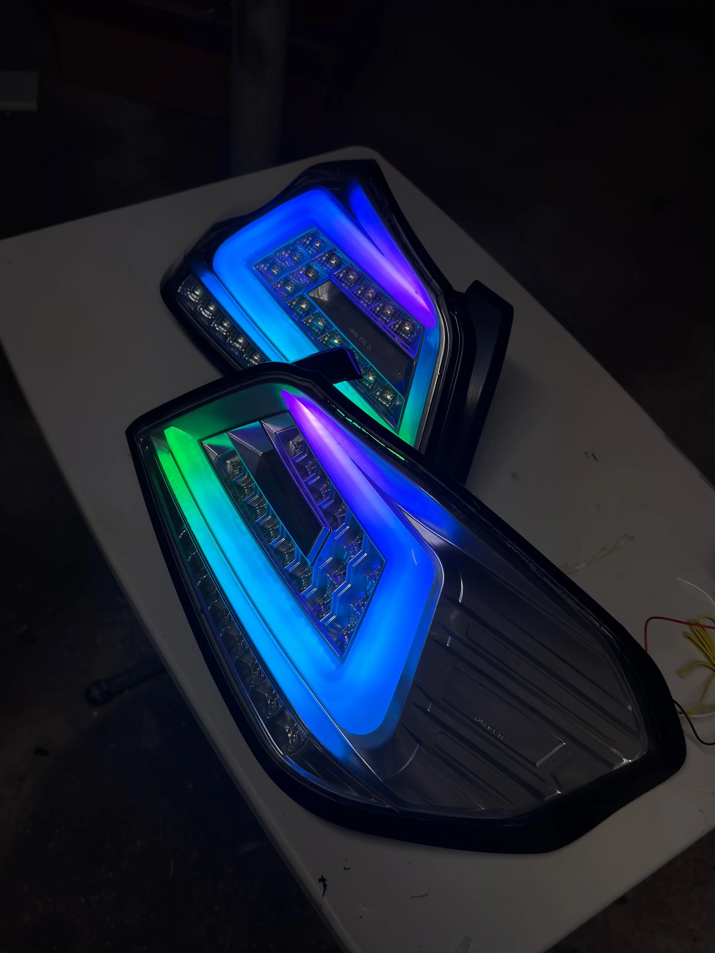 Two LED headlight headlights with colorful LED accents, resting on a white surface in a dark environment.