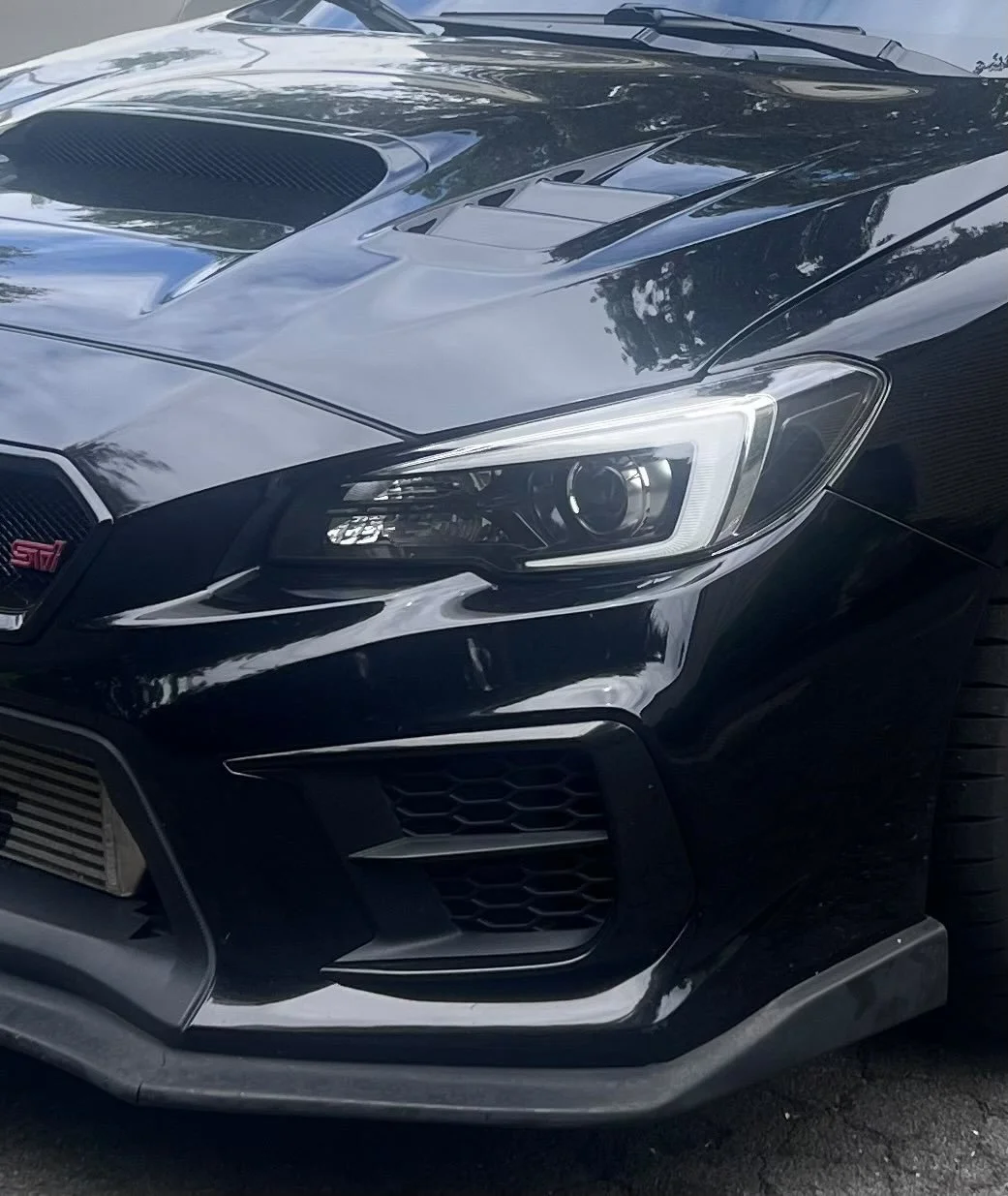 Front view of a black Subaru WRX STI car with a prominent hood scoop, modern headlight design, and grille, parked outdoors with reflections of trees and sky on the car's surface.