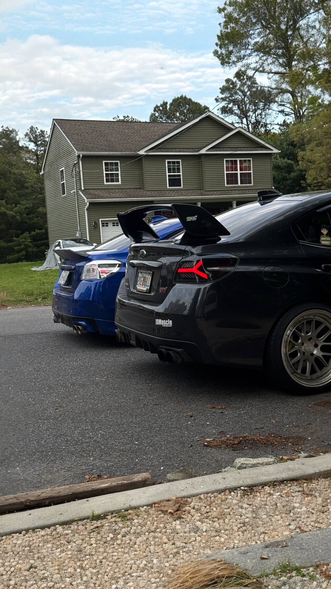 Two sports cars, one black and one blue, parked on a residential street. The black car has a large rear wing and branding stickers, and the house in the background is green with a brown roof.