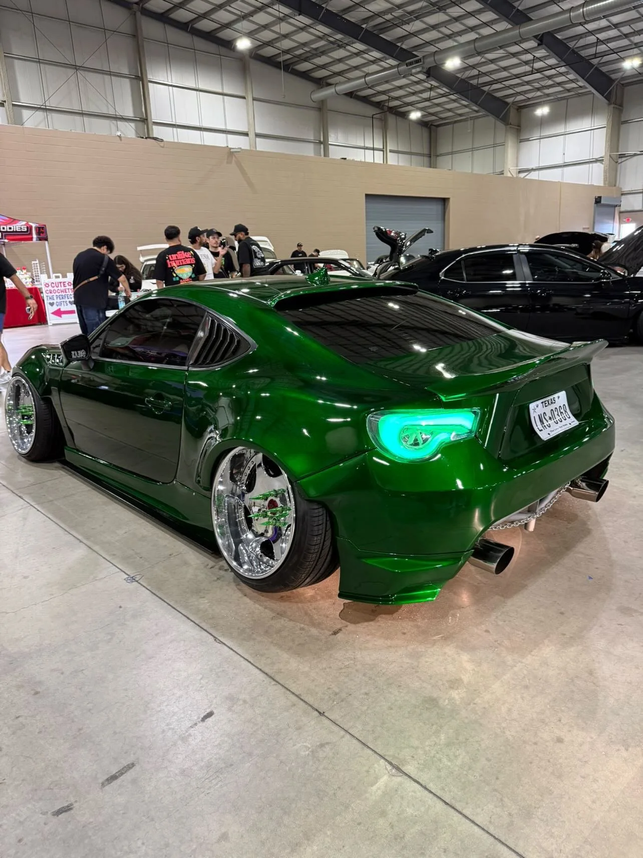 A green modified sports car with wide wheels and custom headlights displayed at an indoor car show, with a group of people in the background.
