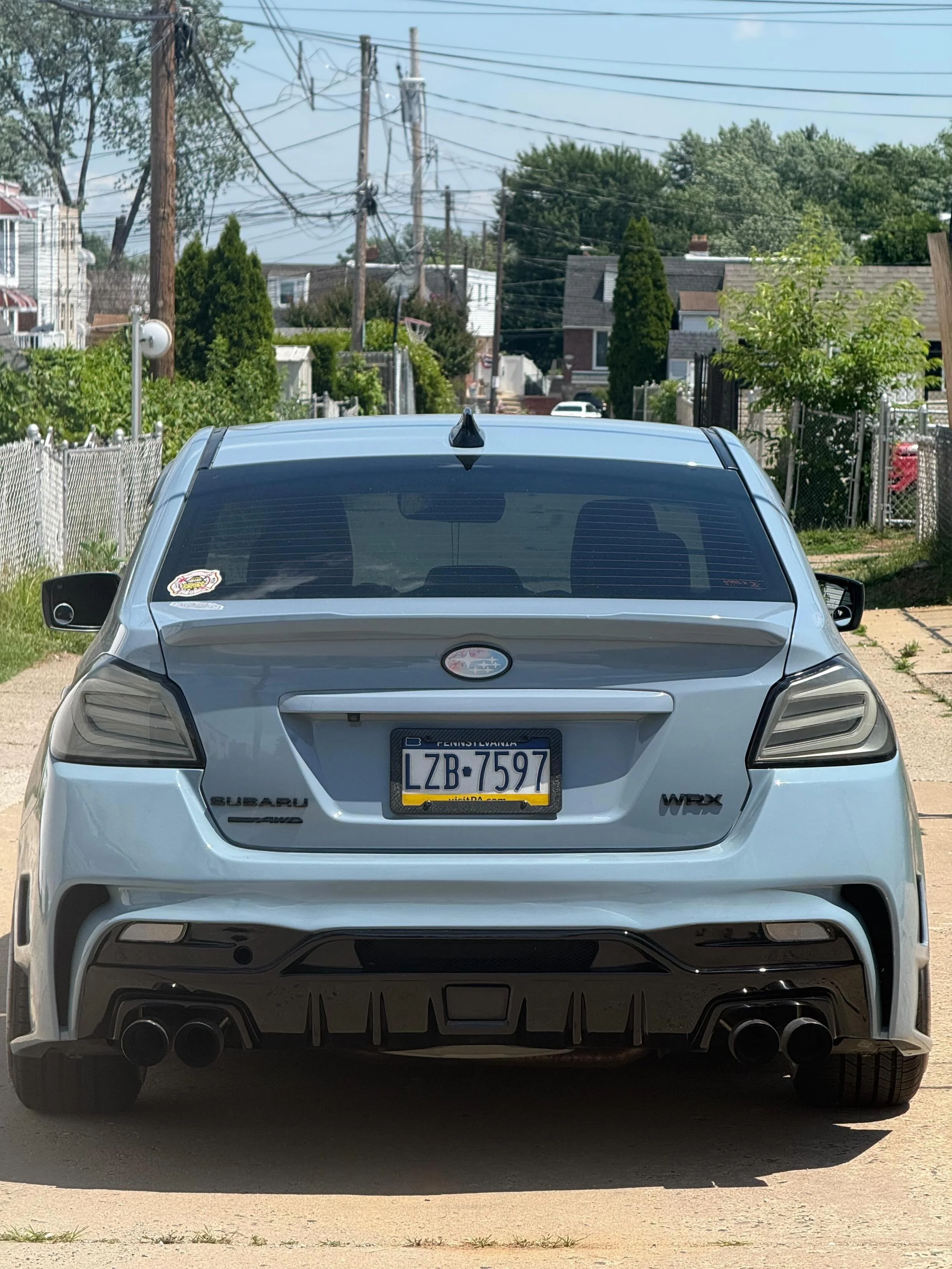 Rear view of a silver Subaru WRX parked on a driveway with houses and trees in the background.