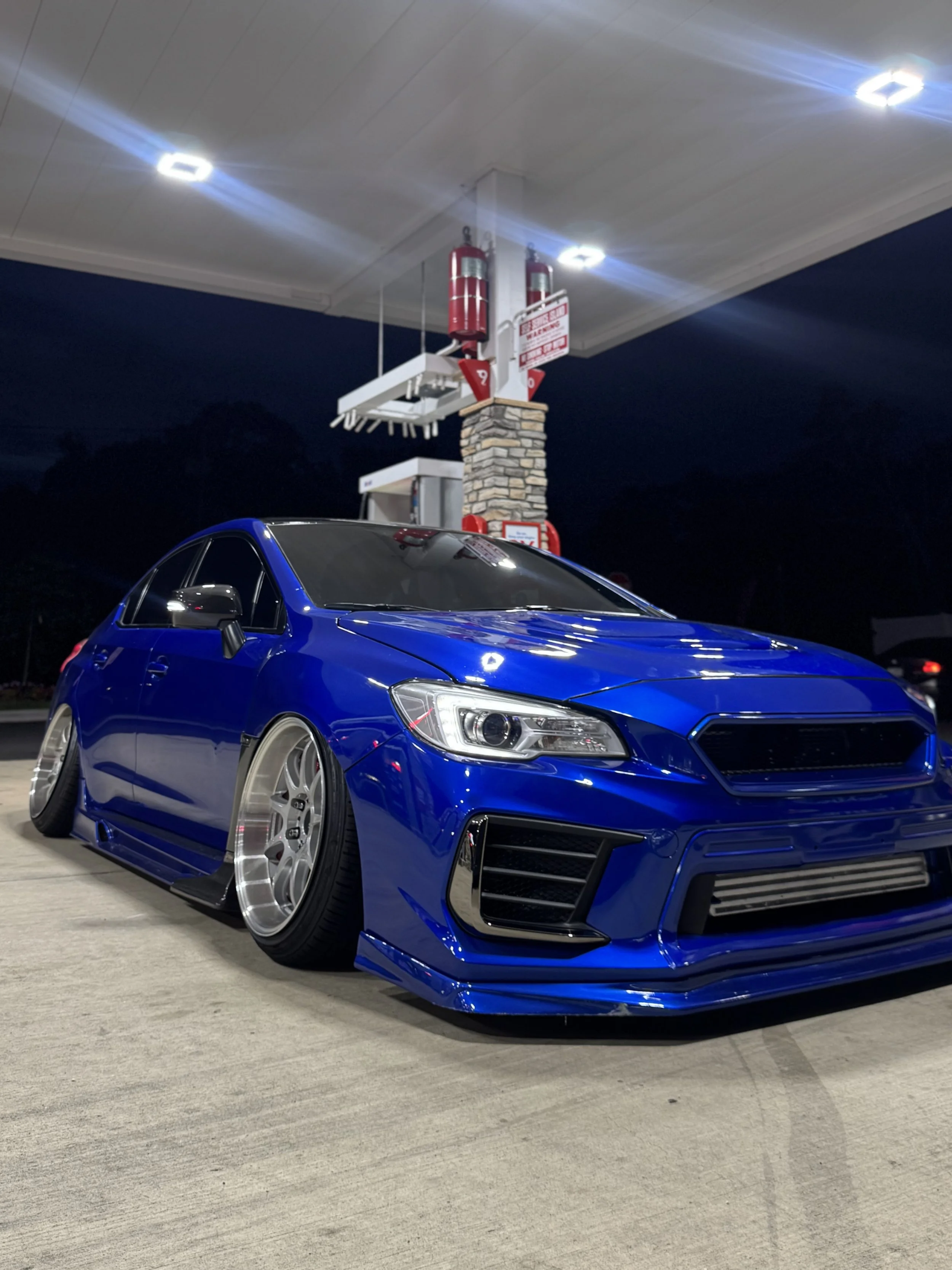 A shiny blue sports car parked on a concrete surface at night, with a gas station in the background illuminated with overhead lights.