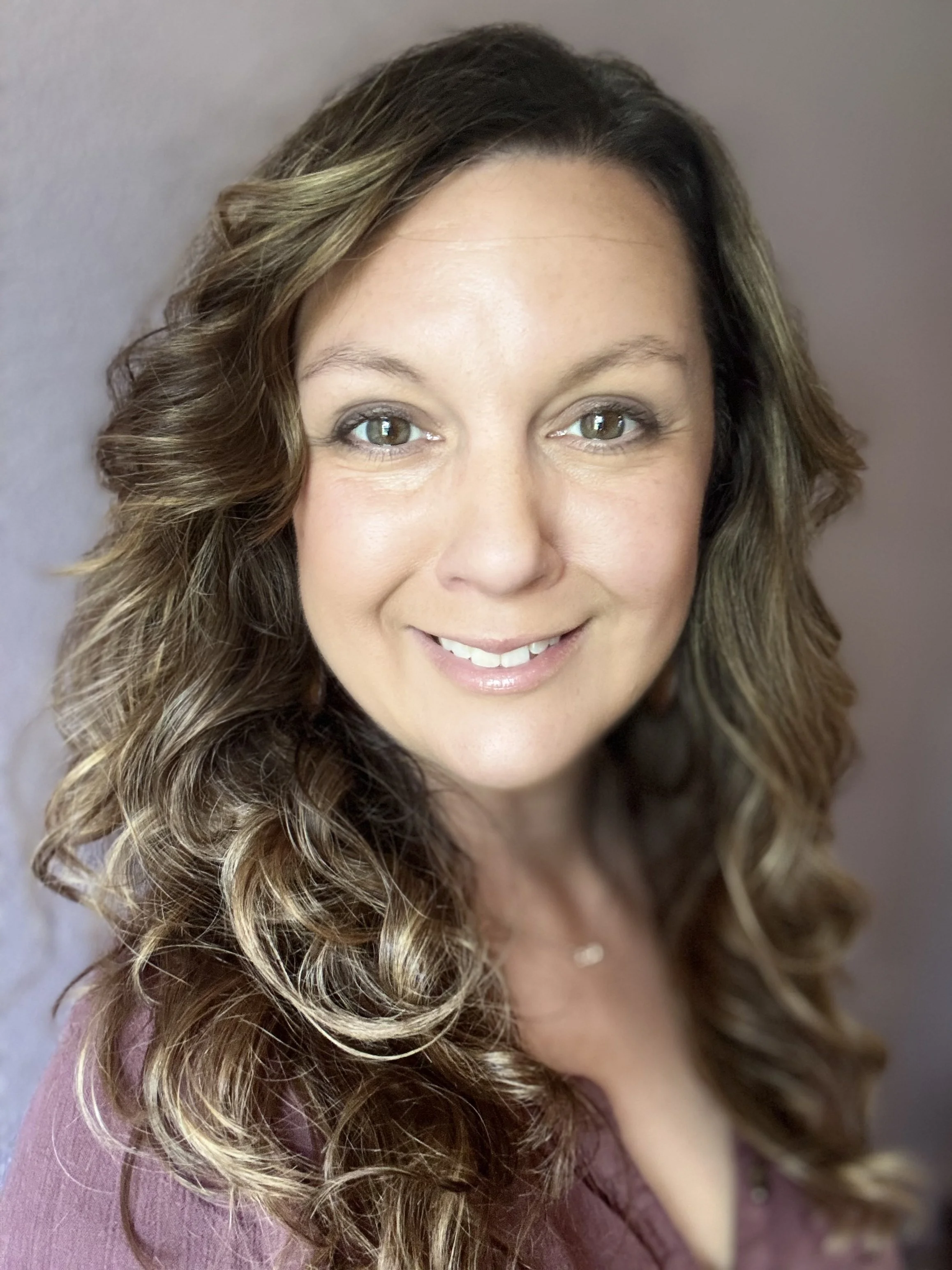 Close-up of a woman with long, wavy hair, smiling and looking at the camera, against a plain background.