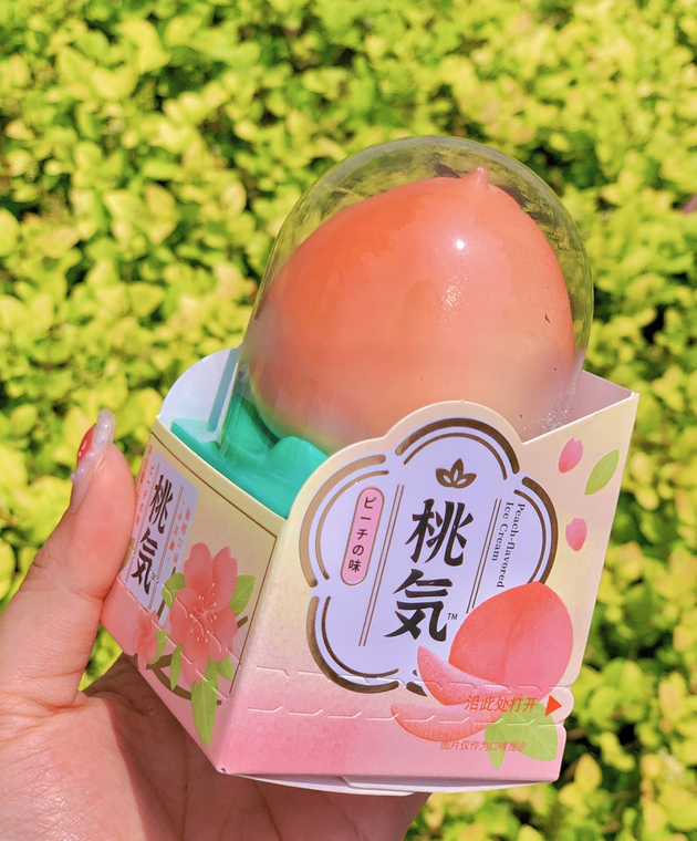 A peach inside a clear plastic container with a decorative pink and white box, held by a person's hand against a background of green foliage.