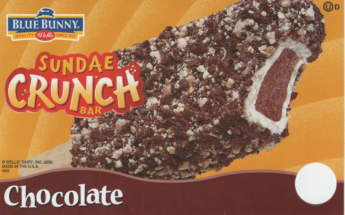 A box of Blue Bunny Sundae Crunch Bar, a chocolate ice cream bar coated with chocolate and crunchy cookie bits, with a partial view of the bar showing chocolate ice cream, a cookie layer, and a chocolate coating.