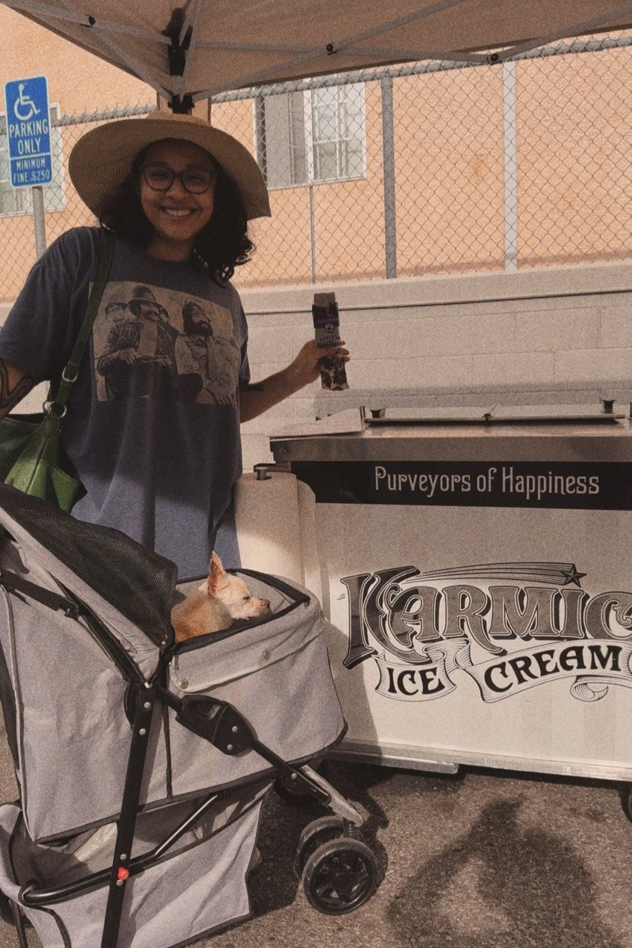 A woman with glasses, a wide-brimmed hat, and a graphic t-shirt stands next to an ice cream cart labeled 'Kermic Ice Cream' with the tagline 'Purveyors of Happiness.' She is holding a small ice cream sandwich in her hand, and a small dog is sitting in a stroller nearby. The backdrop includes a chain-link fence and a building.