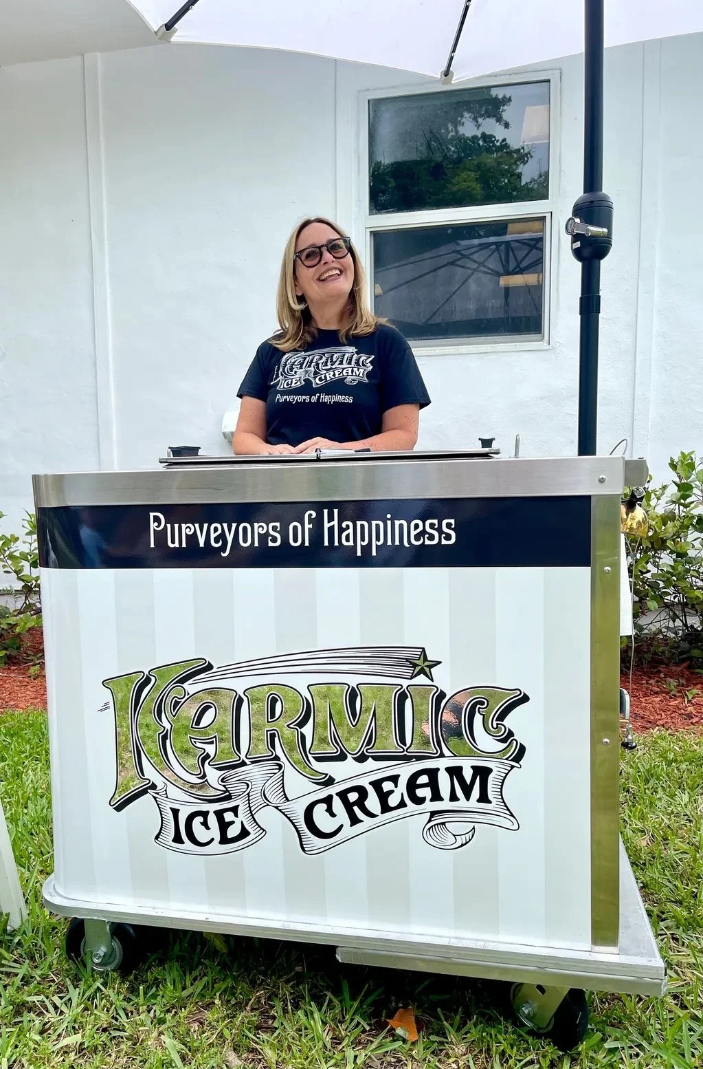 A woman with glasses and blonde hair standing behind a cart that says 'Karmic Ice Cream' and 'Purveyors of Happiness'. She is smiling and wearing a black T-shirt with the same logo. There is a white umbrella and a white building with two windows in the background, along with some green plants and grass.