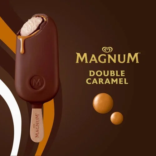 Magnum Double Caramel ice cream bar with a bite taken out, showing caramel and ice cream inside, on a brown background with caramel-colored spheres and the Magnum logo.