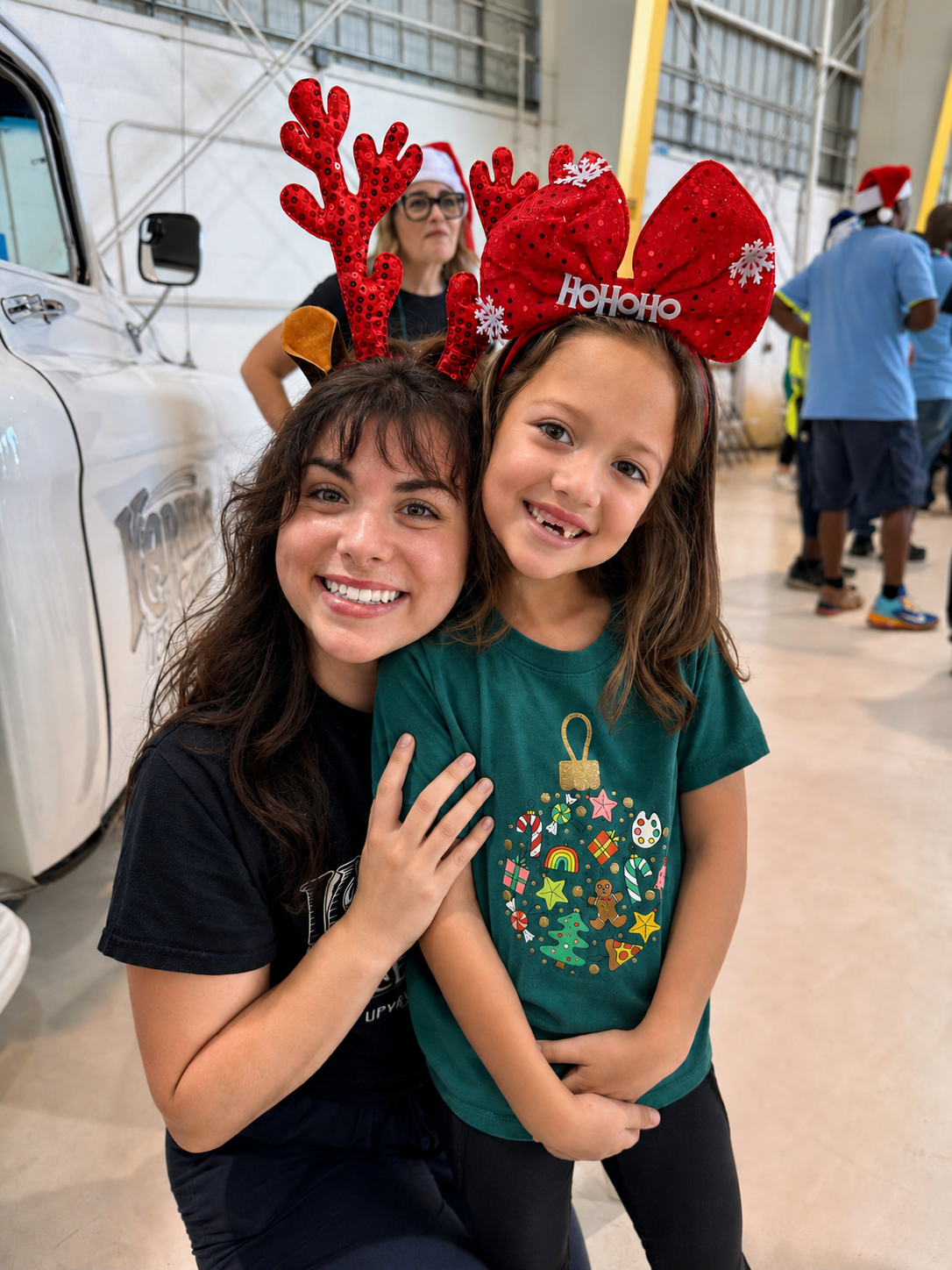 Two smiling girls, one older and one younger, celebrating Christmas indoors. The older girl is wearing reindeer antlers, and the younger is wearing a large red bow headband with white snowflakes and a 'Ho Ho Ho' sign. People are in the background, some in Santa hats.