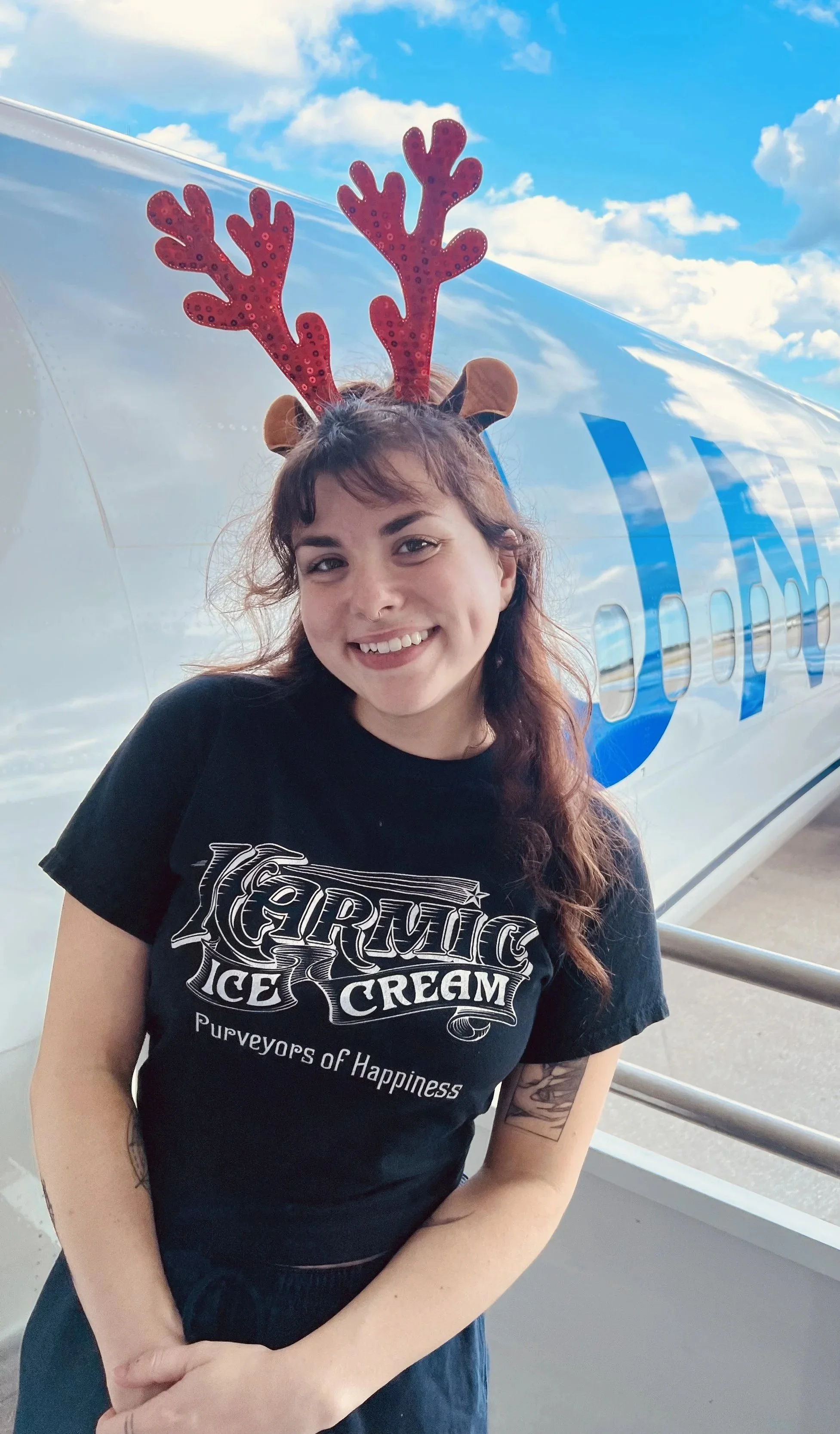 A woman with long brown hair wearing reindeer antlers and a black T-shirt with 'Germic Ice Cream' logo, standing outdoors near an aircraft with blue markings and a partly cloudy sky in the background.