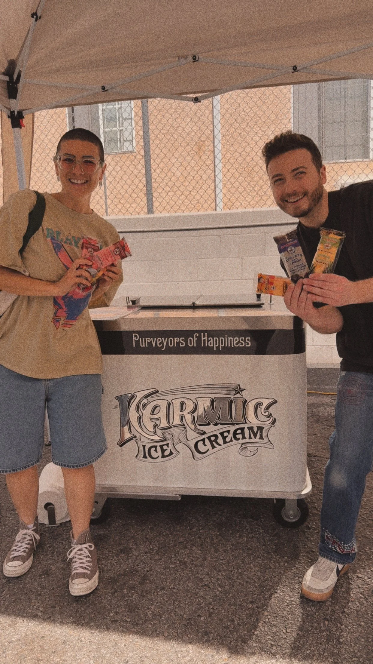 Two smiling people at an ice cream stand, holding packaged ice cream bars, standing beside a cart with a sign that reads 'Karmic Ice Cream,' outdoors under a canopy, with a chain-link fence and building in the background.