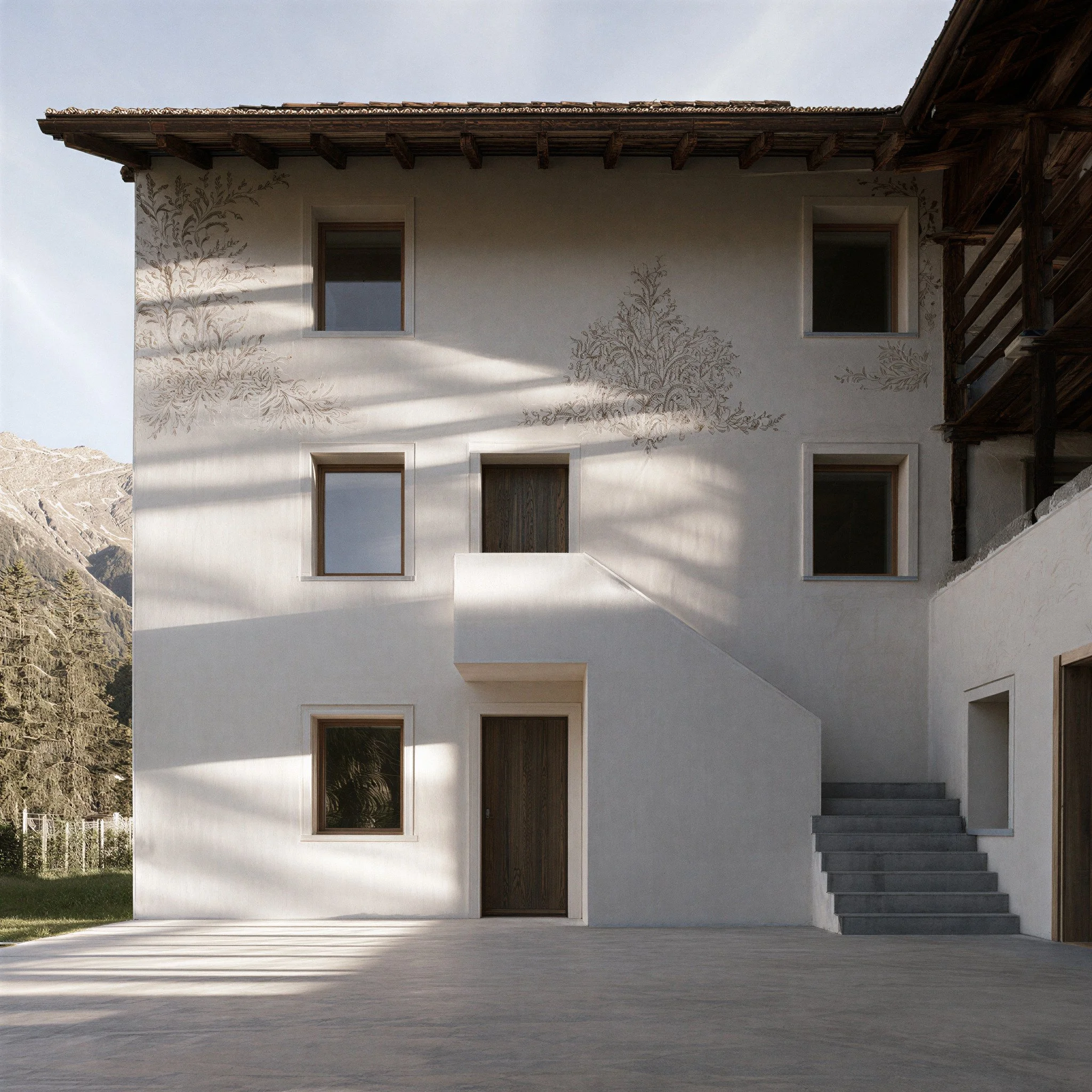 Modern white building with multiple rectangular windows and decorative tree-branch illustrations on the facade. Stairs are visible on the right side, leading to an upper level. Mountain landscape in the background.