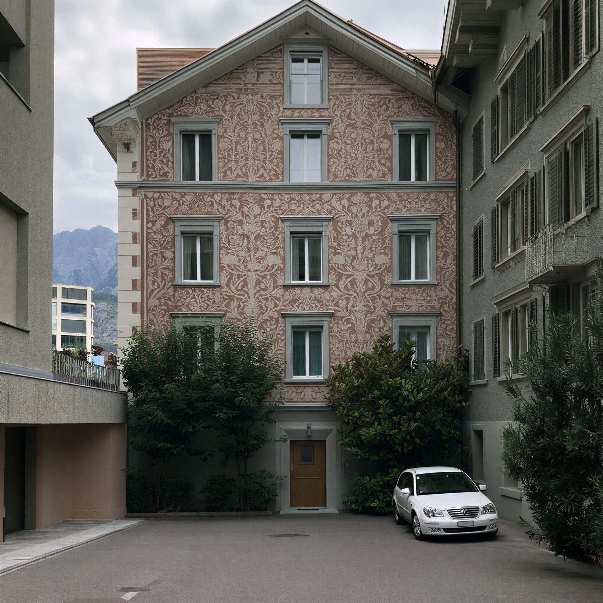 A multi-story building with a distinctive pinkish facade decorated with floral patterns, surrounded by other buildings, a parked silver car, and mountains in the background under a cloudy sky.