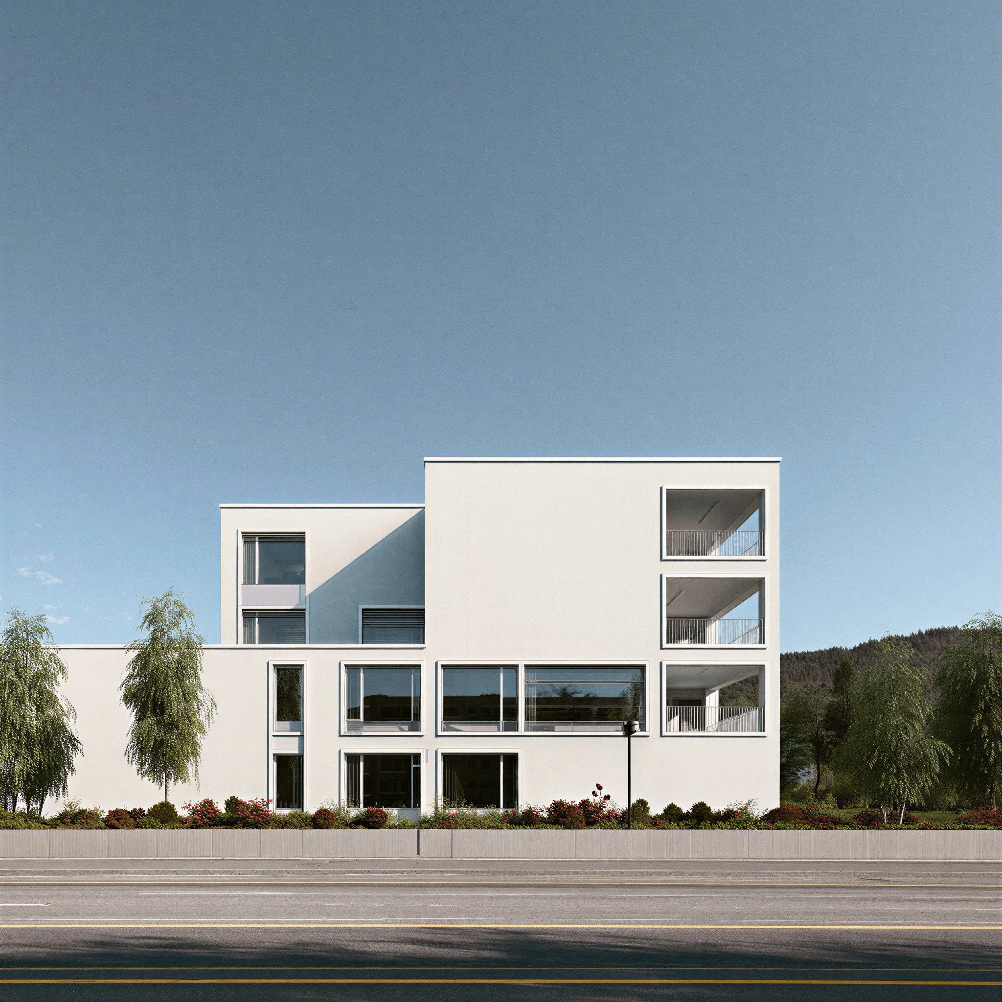 Modern white building with multiple balconies and large windows, landscaped with trees and bushes, set against a clear blue sky.