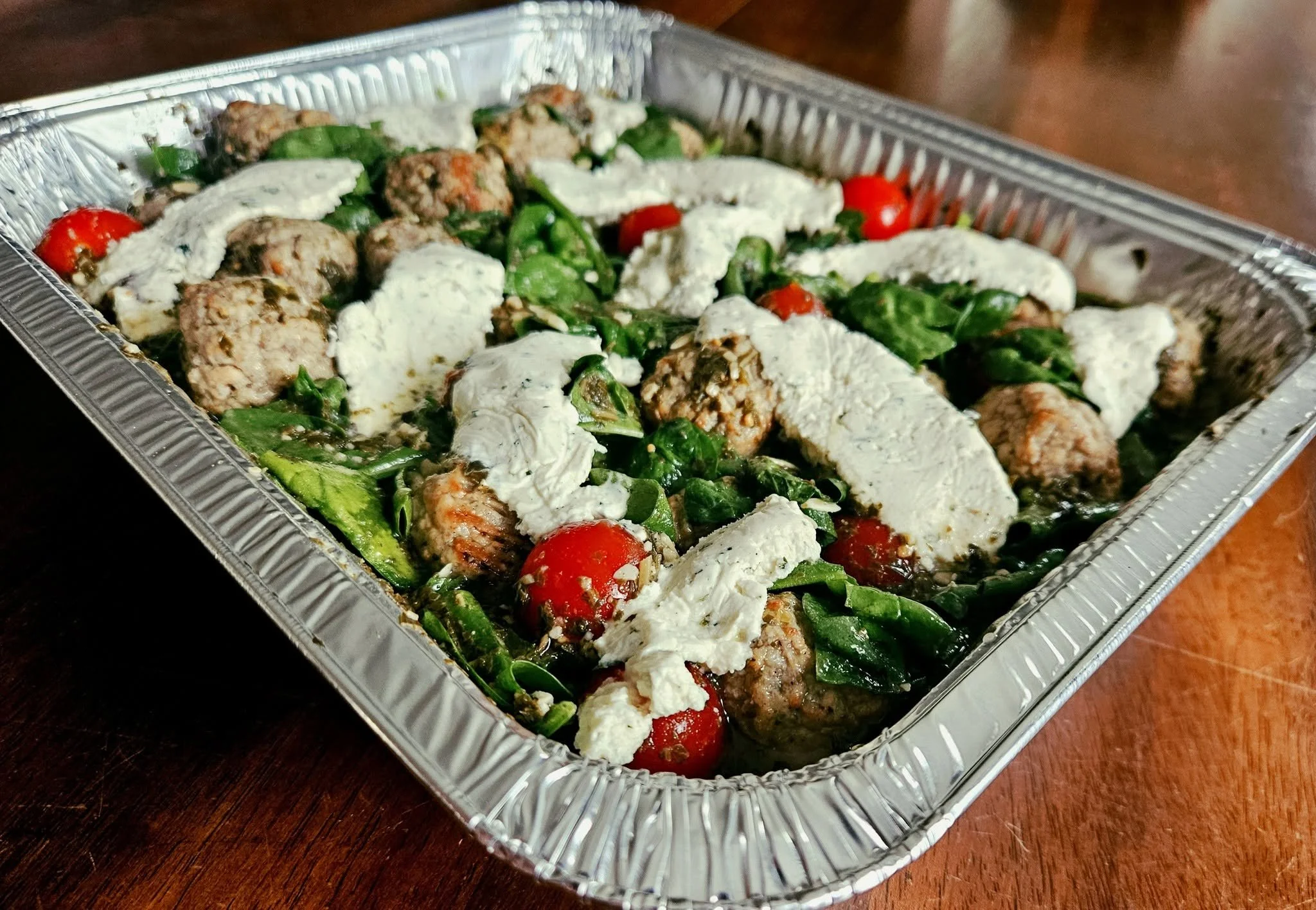 A takeout aluminum tray holding cooked meatballs, spinach, cherry tomatoes, and dollops of white sauce.