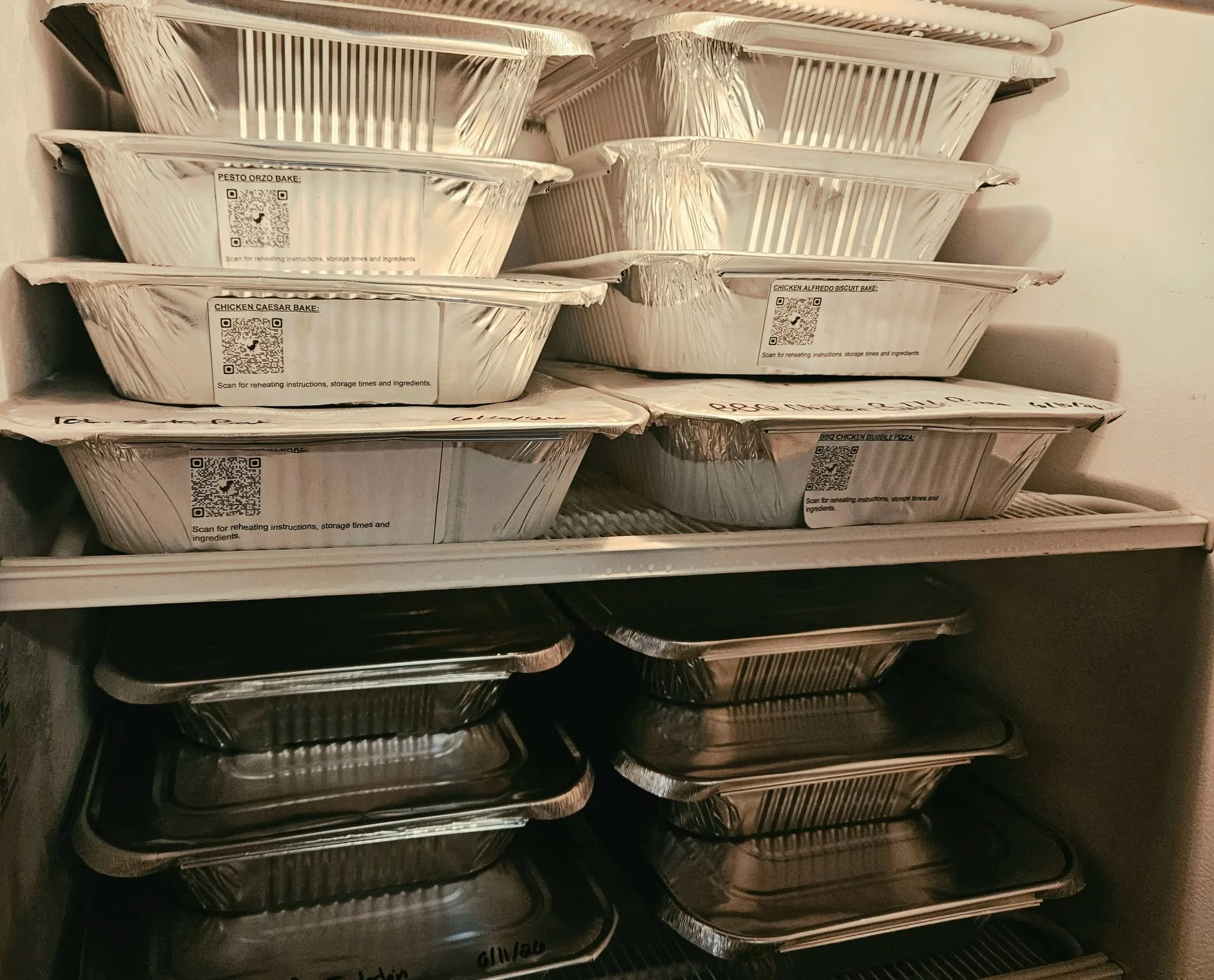 A collection of aluminum foil food containers on a shelf, labeled for baked dishes such as pesto orzo, chicken Caesar, chicken Alfredo, and BBQ chicken bubble pizza.