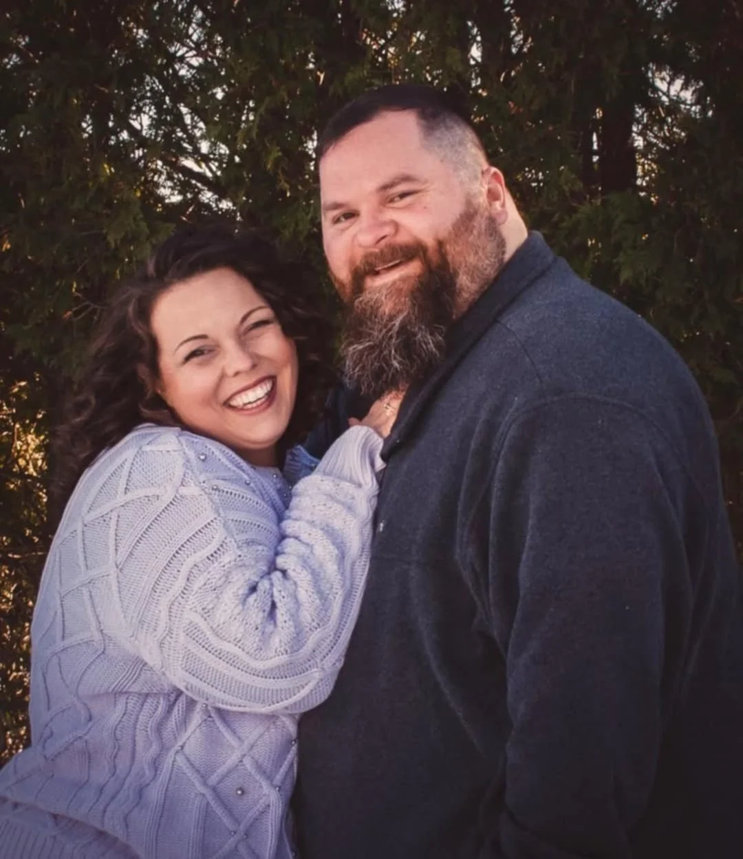 A happy woman with curly dark hair and a white sweater, and a man with a beard and dark jacket, smiling and posing together outdoors in front of trees.