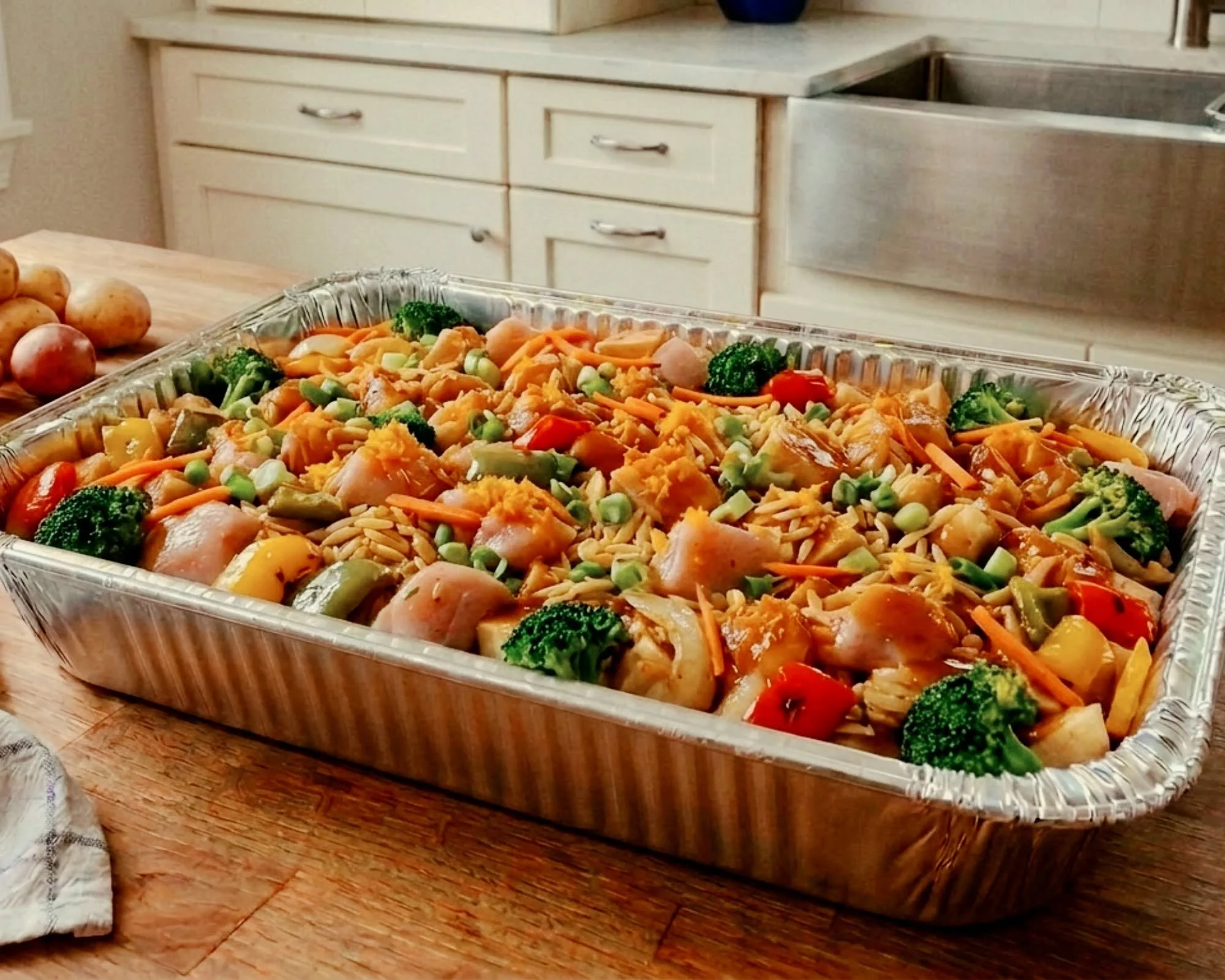 A large aluminum tray filled with a colorful vegetable casserole containing broccoli, carrots, peas, mushrooms, and other vegetables. The tray is on a wooden table next to a small pile of potatoes. In the background, there are cream-colored kitchen cabinets and a stainless steel sink.