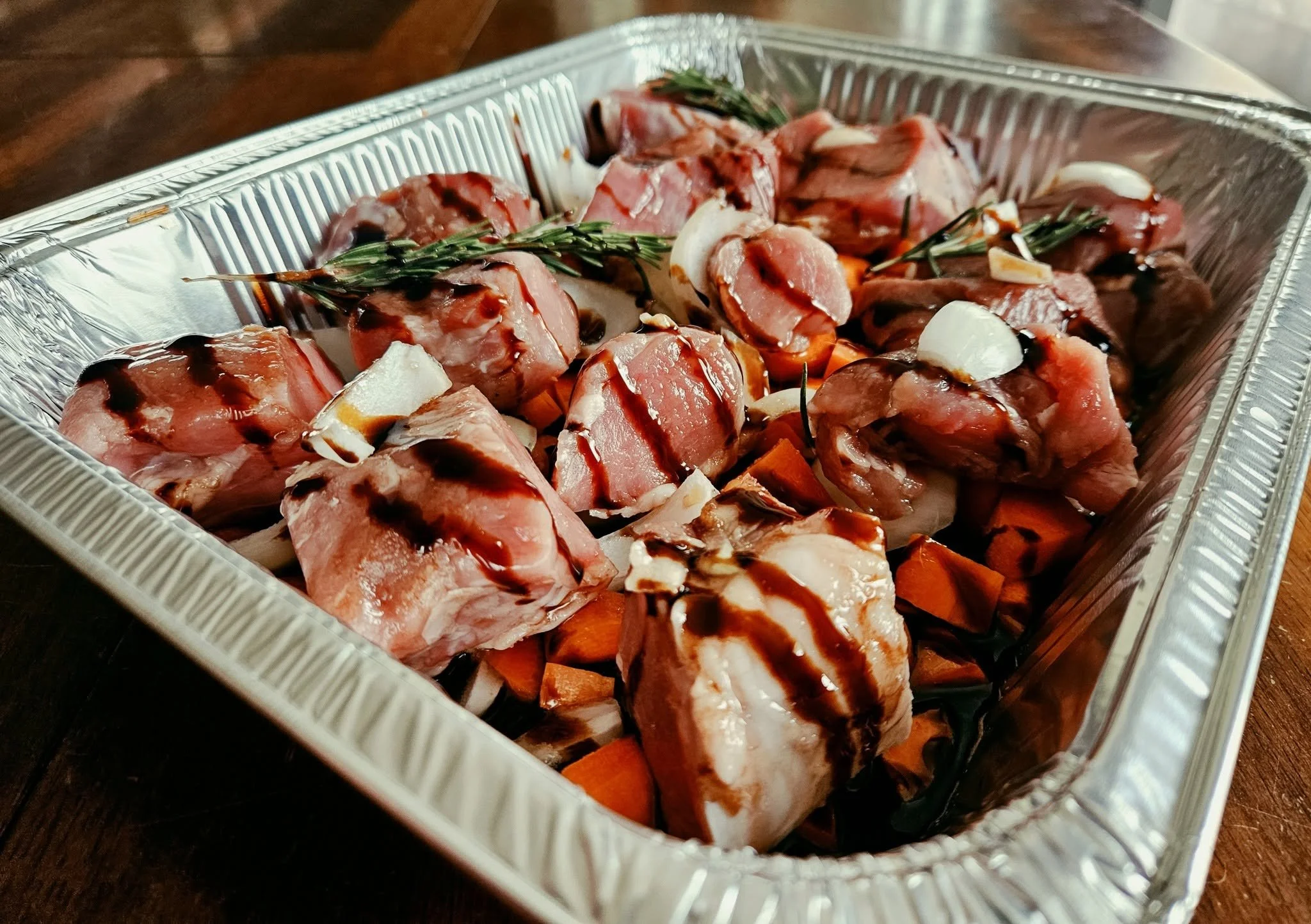 Raw lamb chops garnished with rosemary and garlic in a foil-covered container on a wooden surface.