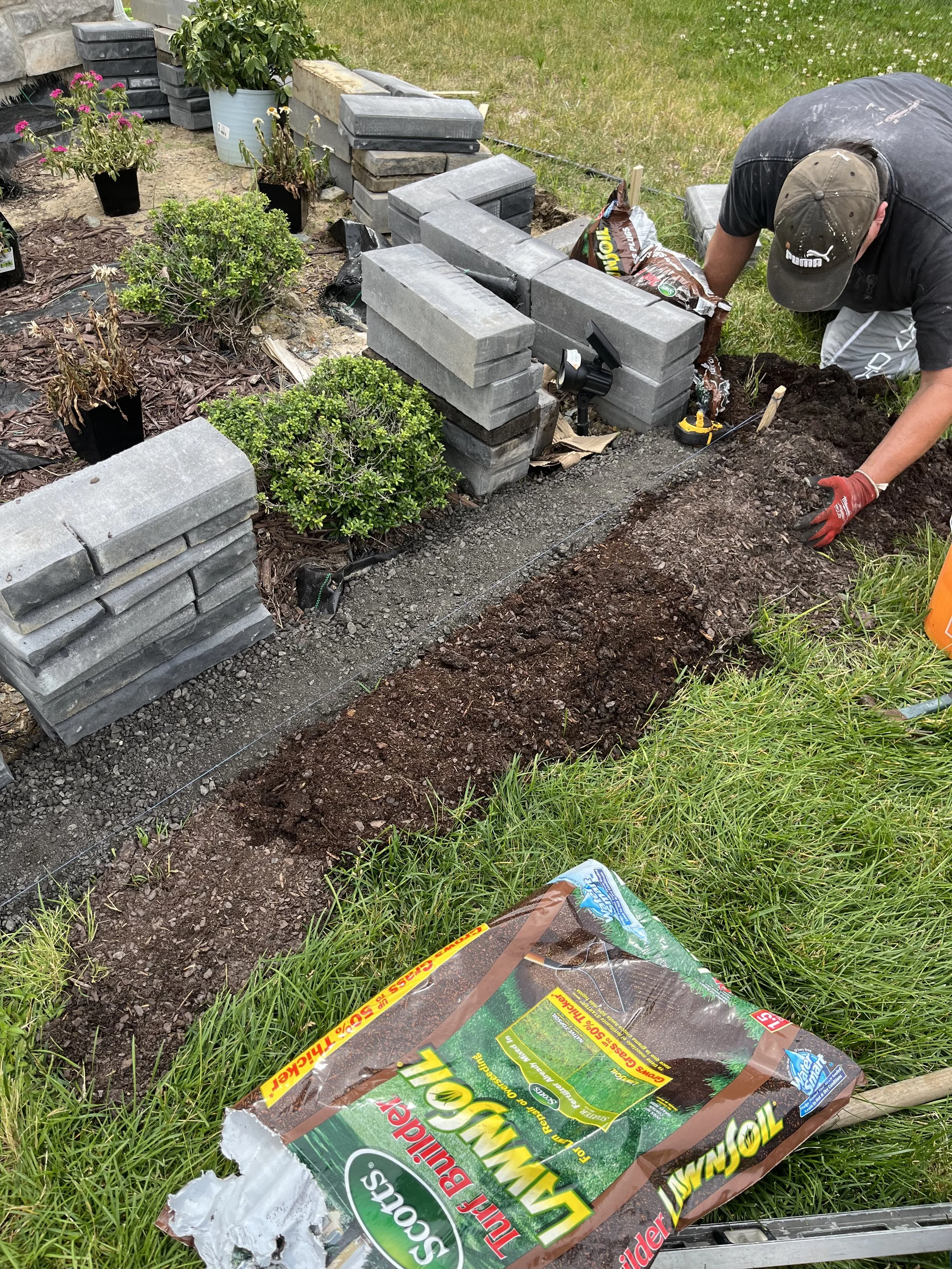A person working on a garden border, laying down bricks and installing landscape fabric. Various potted plants are nearby, and a bag of soil or mulch is on the grass.