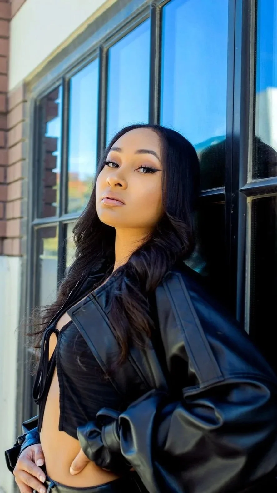 A woman with long black hair leaning against a window with blue-tinted glass, wearing a black leather jacket.