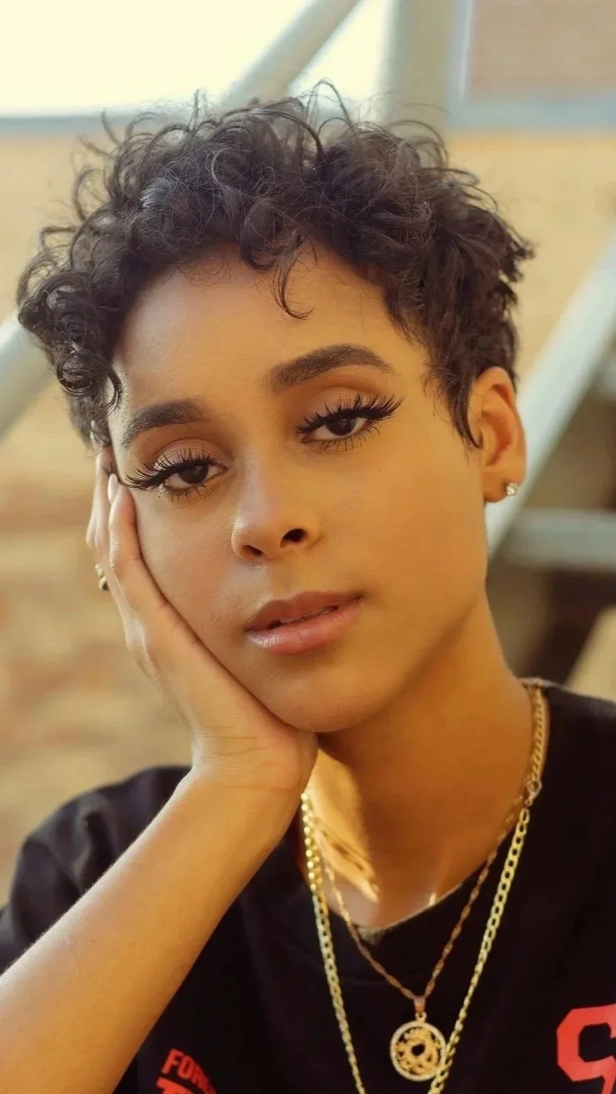 A young woman with short curly hair resting her head on her hand, wearing makeup, earrings, and layered gold necklaces, in an indoor setting.