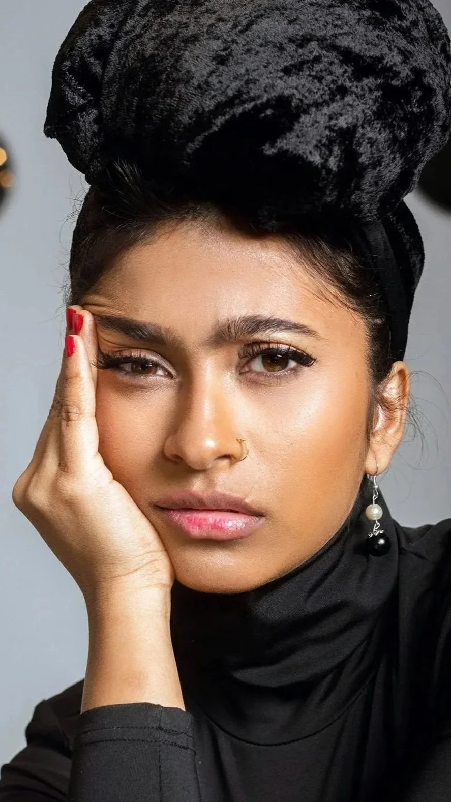 A woman with dark hair wearing a black headwrap, black turtleneck, and pearl earrings, resting her face on her hand, partially winking at the camera.