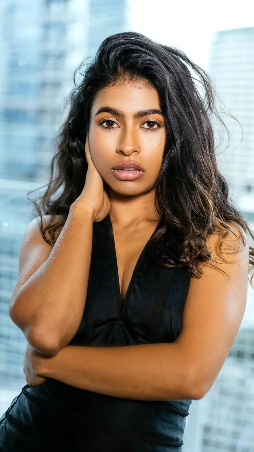 A young woman with long wavy dark hair, wearing a black sleeveless top, posing indoors with a cityscape of tall glass buildings in the background.