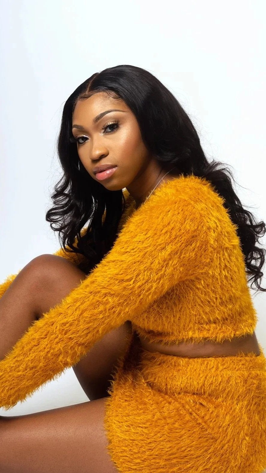 A young woman with black, wavy hair, wearing makeup, and a yellow fuzzy sweater, sitting with her knee up against a plain white background.