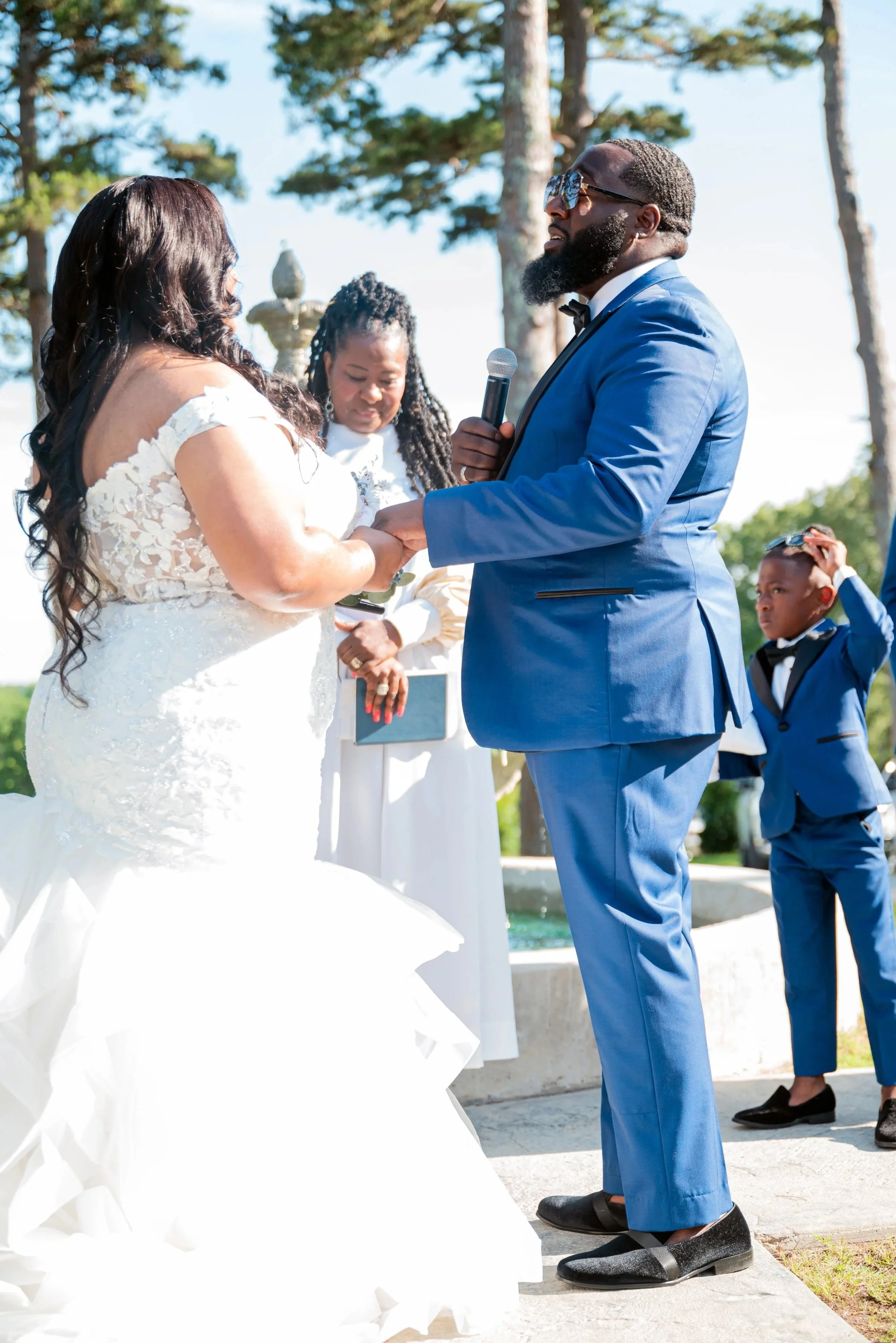A wedding ceremony outdoors with a couple exchanging vows, officiant holding a microphone, and a boy in a blue suit visible in background.