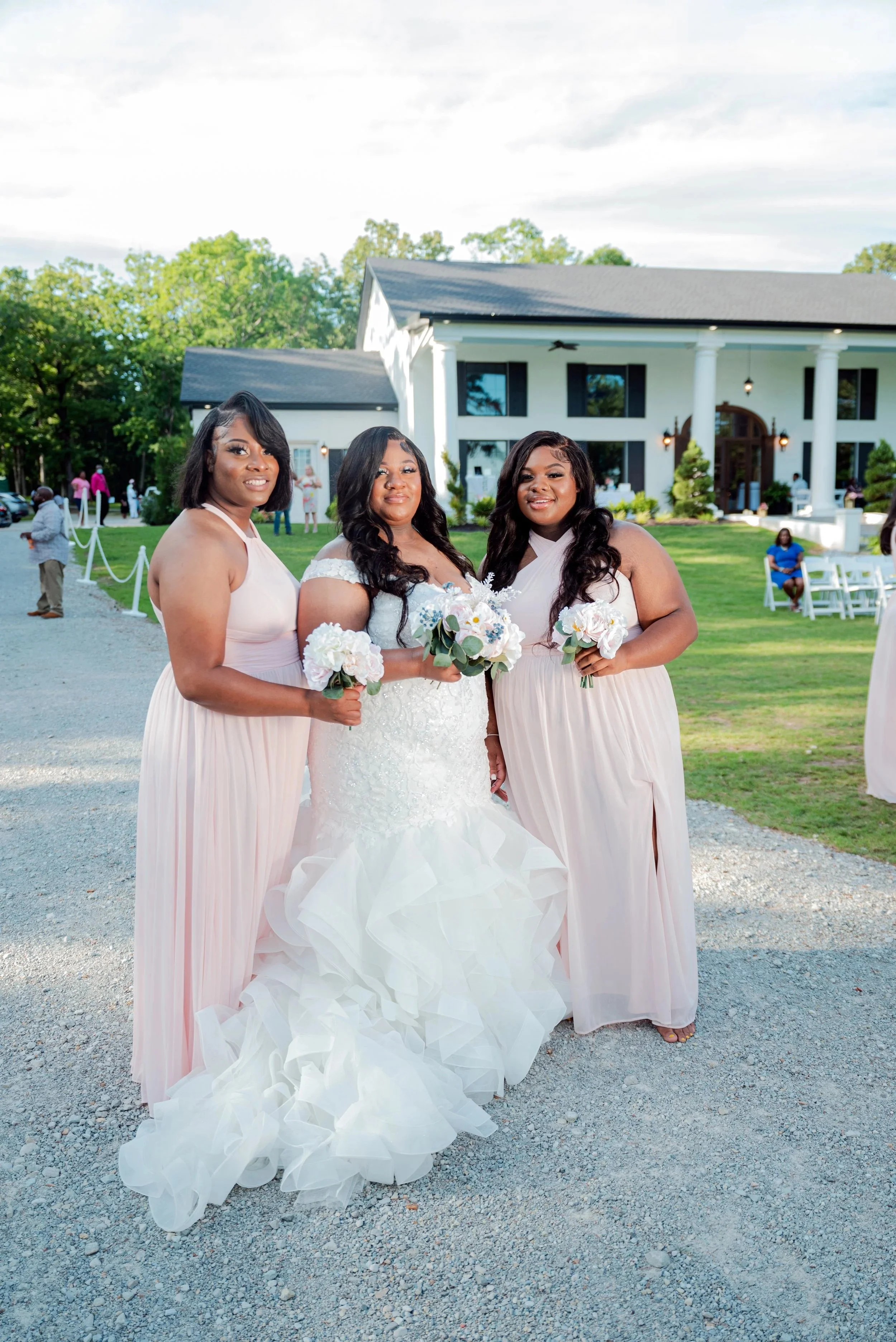 Three women in bridesmaid dresses and a bride in a wedding dress standing outdoors in front of a large house with white columns, holding bouquets of flowers, with guests in the background.