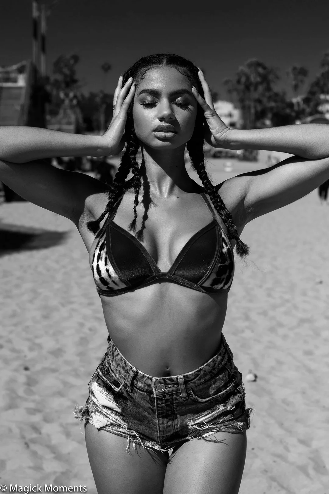 A woman in a leopard print bikini top and ripped denim shorts stands on a beach with her eyes closed, hands on her head, and a relaxed expression. In black and white photograph.