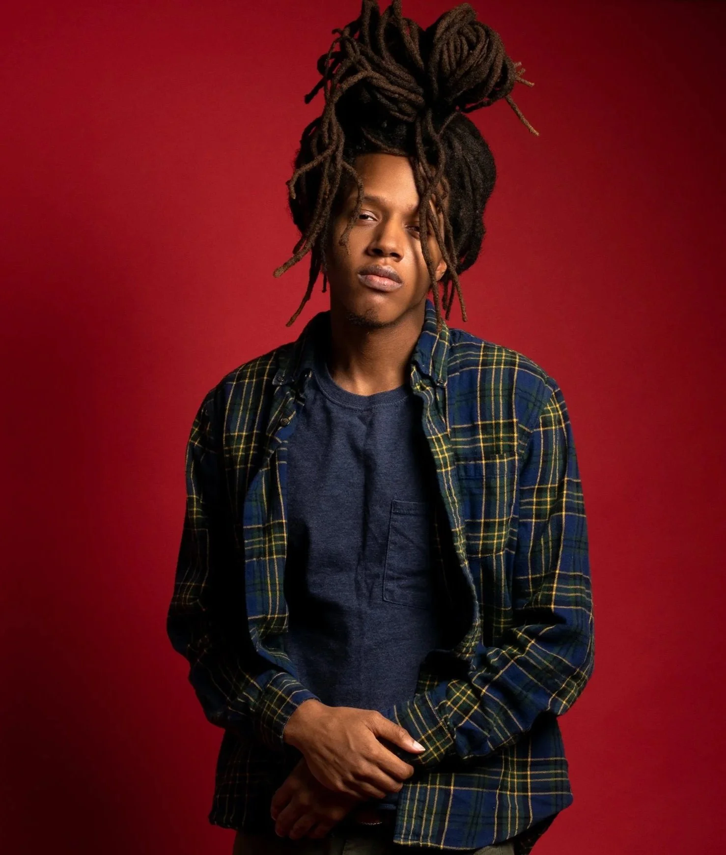 Young man with long dreadlocks, wearing a navy blue t-shirt and plaid shirt, standing against a red background.