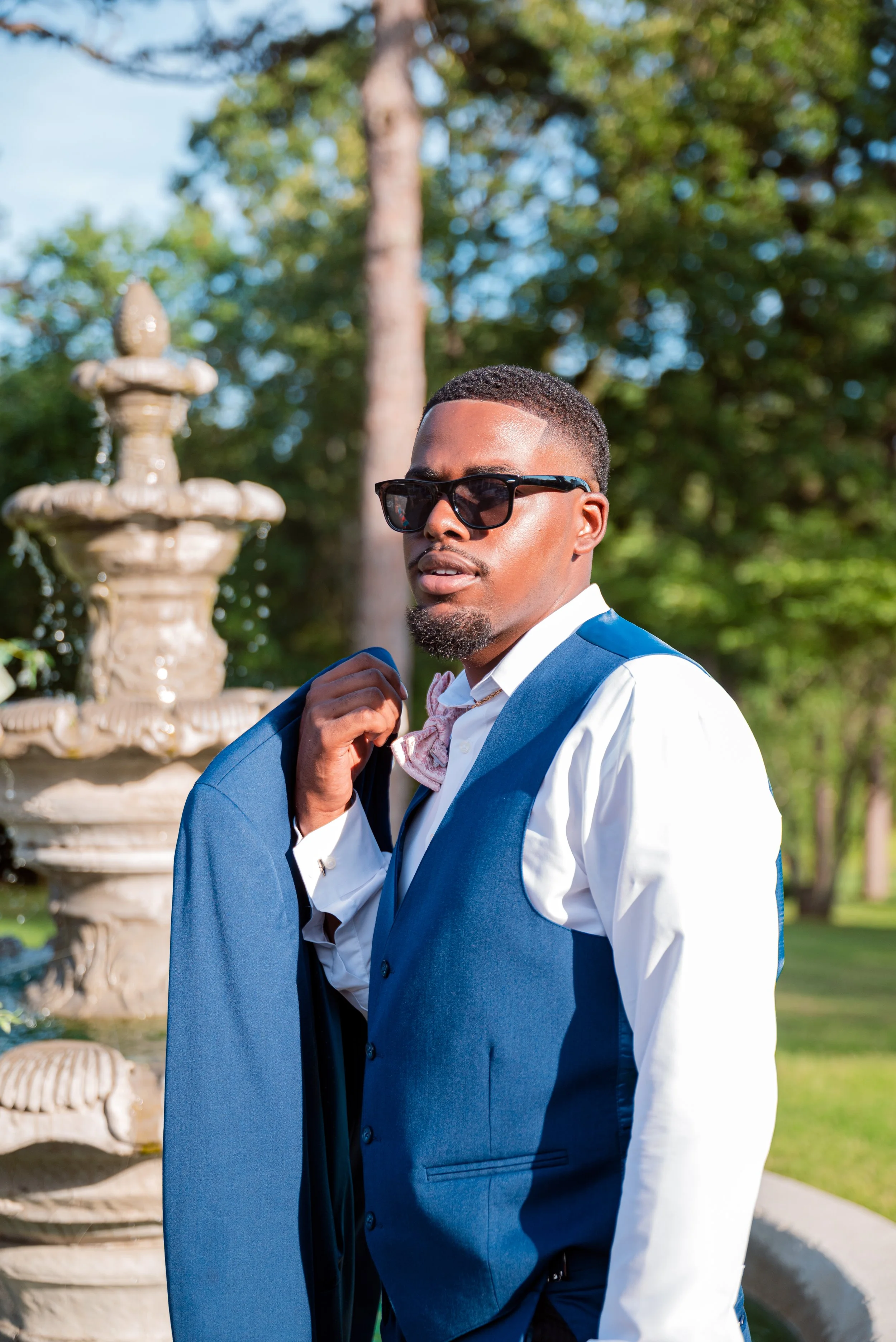 A man with short hair, sunglasses, and a goatee, wearing a white shirt and a blue vest, stands outside near a fountain on a sunny day. He is holding a jacket over his shoulder.