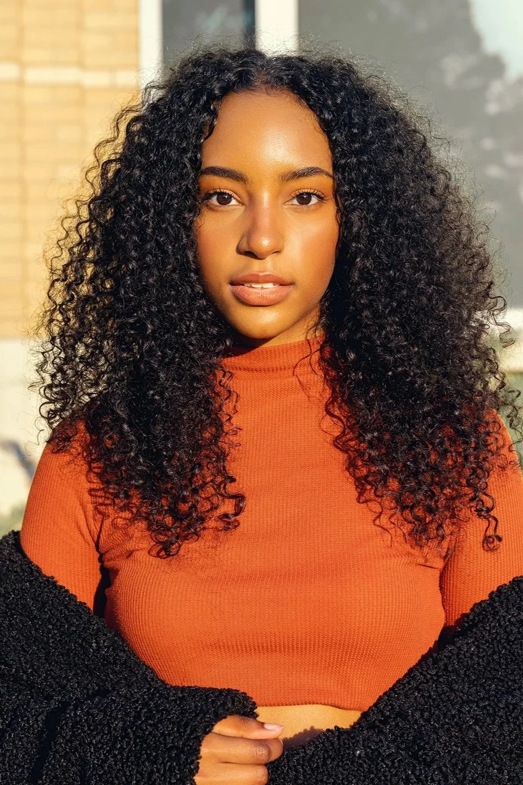 A young woman with dark, curly hair, wearing an orange top and a black cardigan, standing outdoors in bright sunlight.