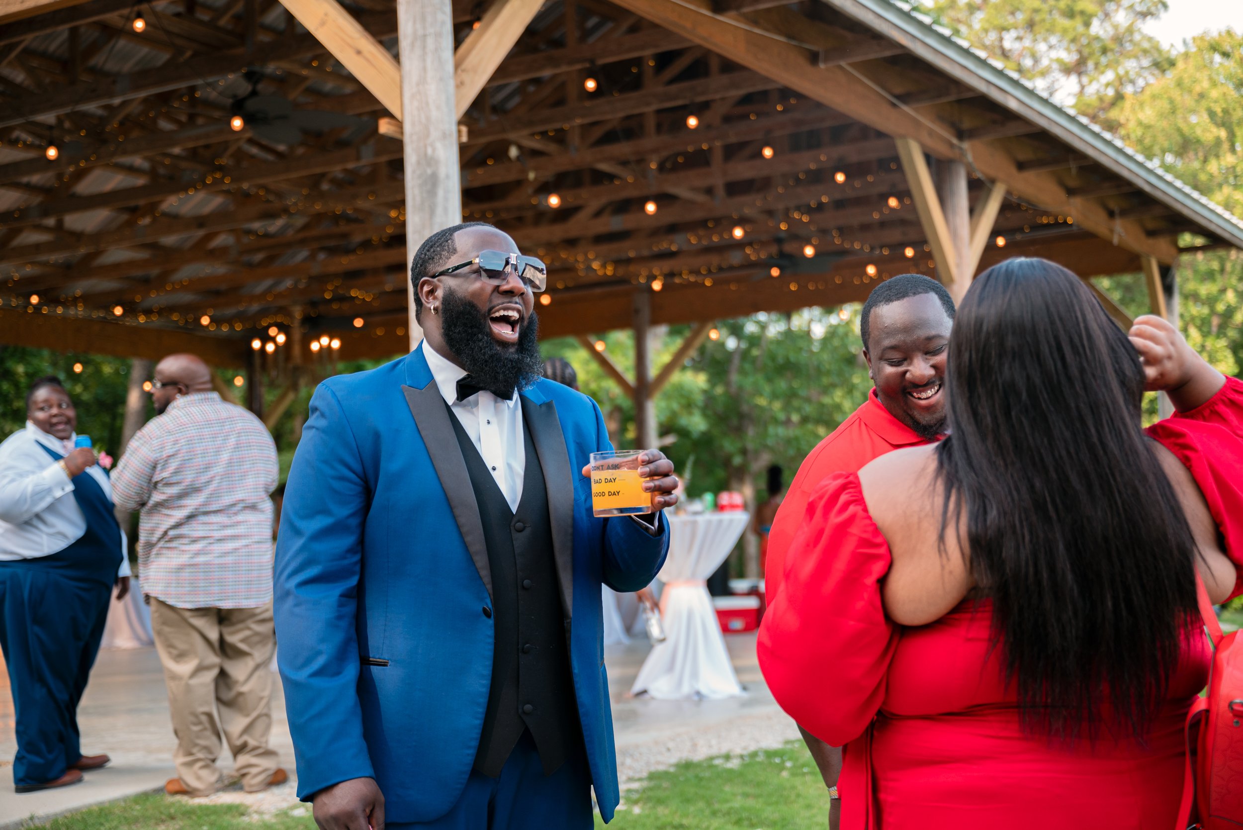 Man in a blue suit with sunglasses and a beard holding a drink, laughing and socializing at a gathering outdoors under a wooden pavilion with string lights. Other people are in the background, some chatting and smiling.